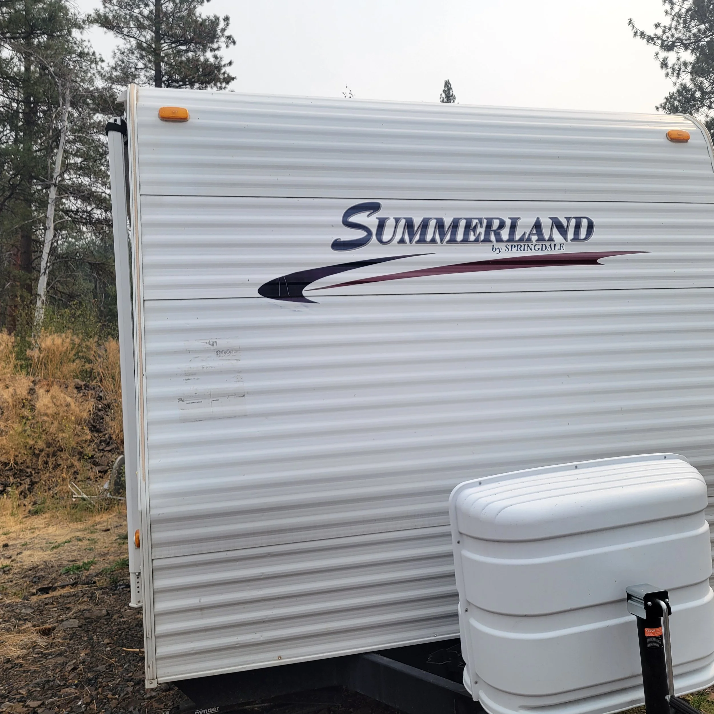 A white travel trailer with the brand name 'Summerland by Springdale' on the side, parked outdoors with trees and a dirt ground in the background.