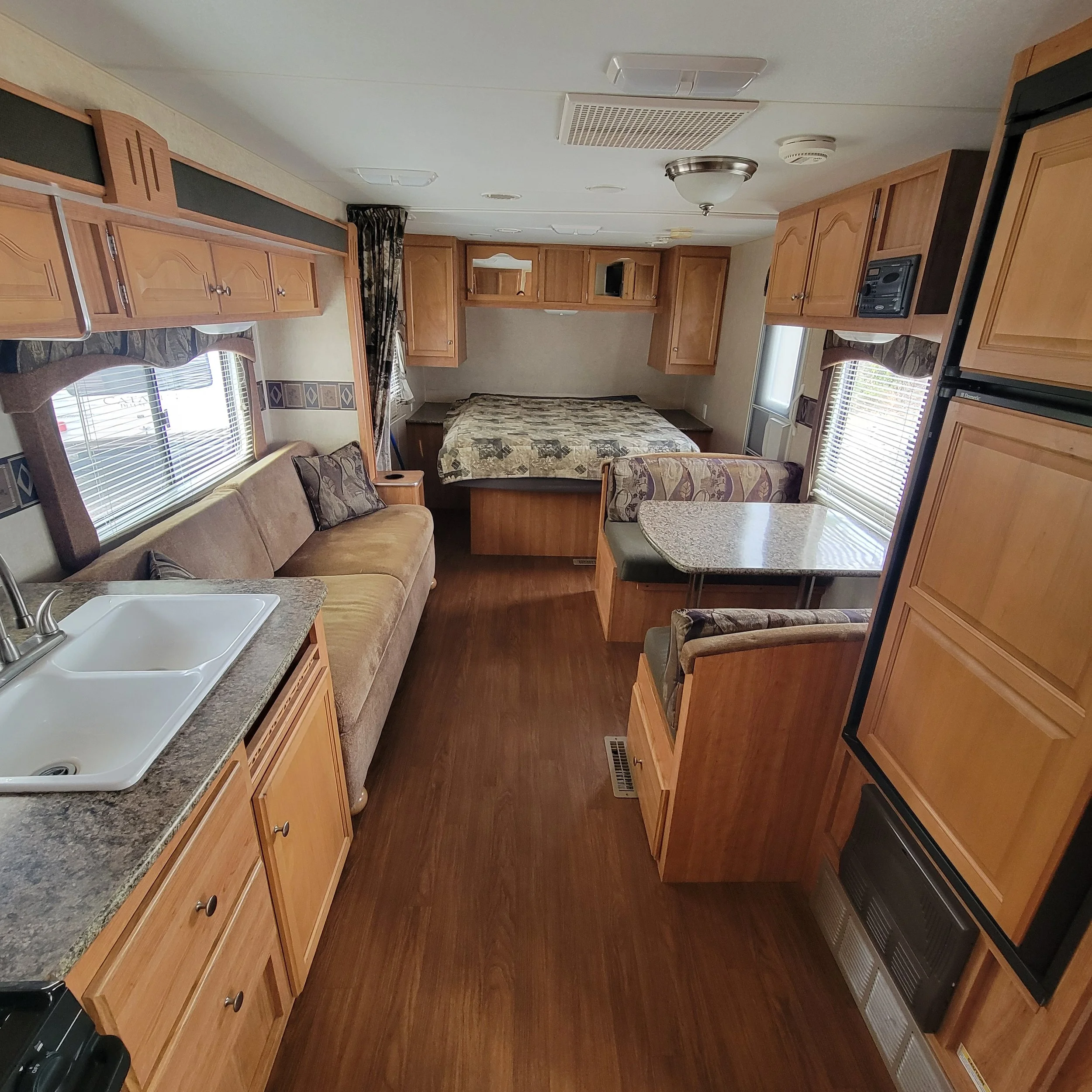 Interior of a compact RV with wooden cabinets, a sofa, a dining table, a bed, and a kitchenette.