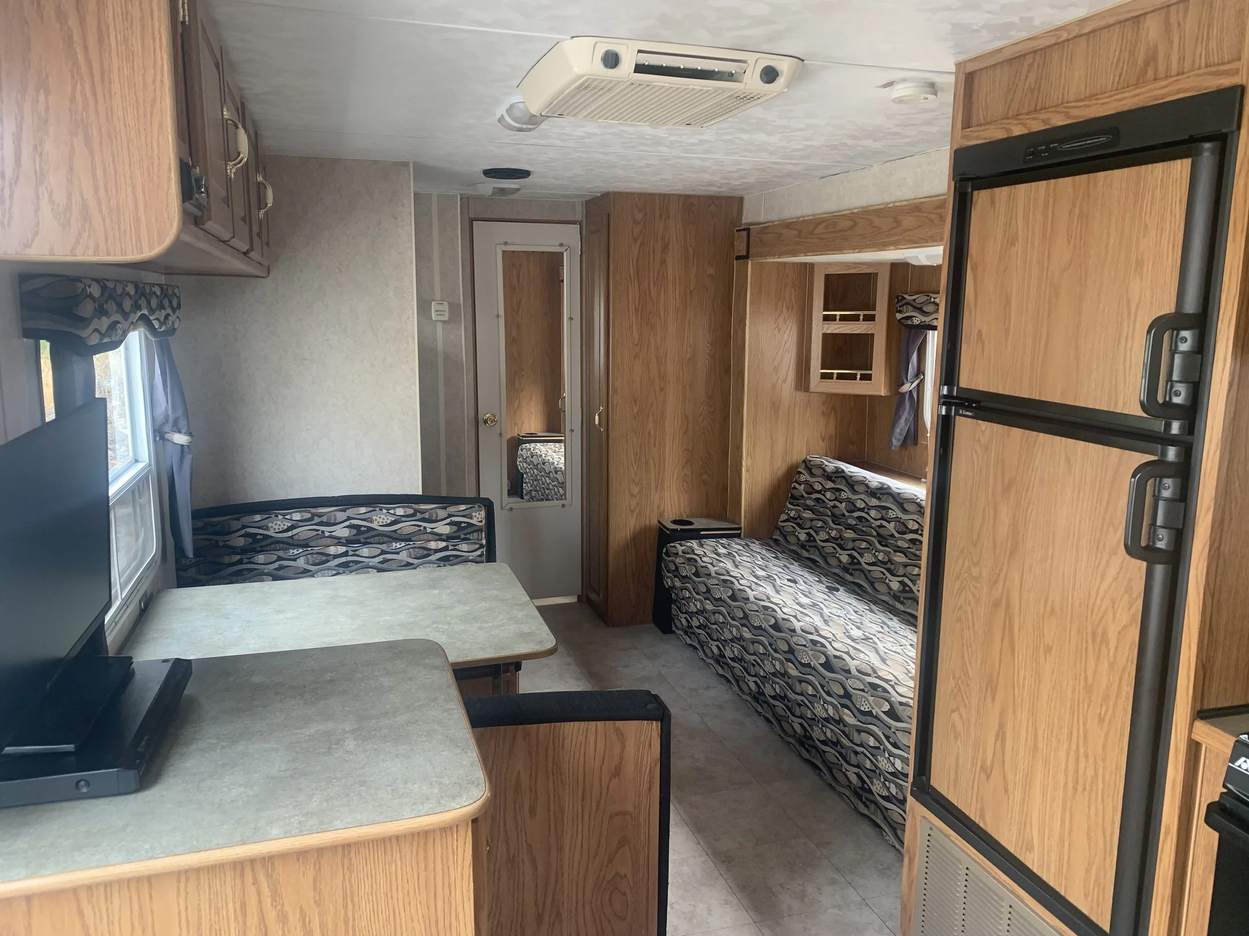 Interior of a small mobile home or RV with wooden cabinets, a small dining table, a sofa bed, a refrigerator, and a window with curtains.