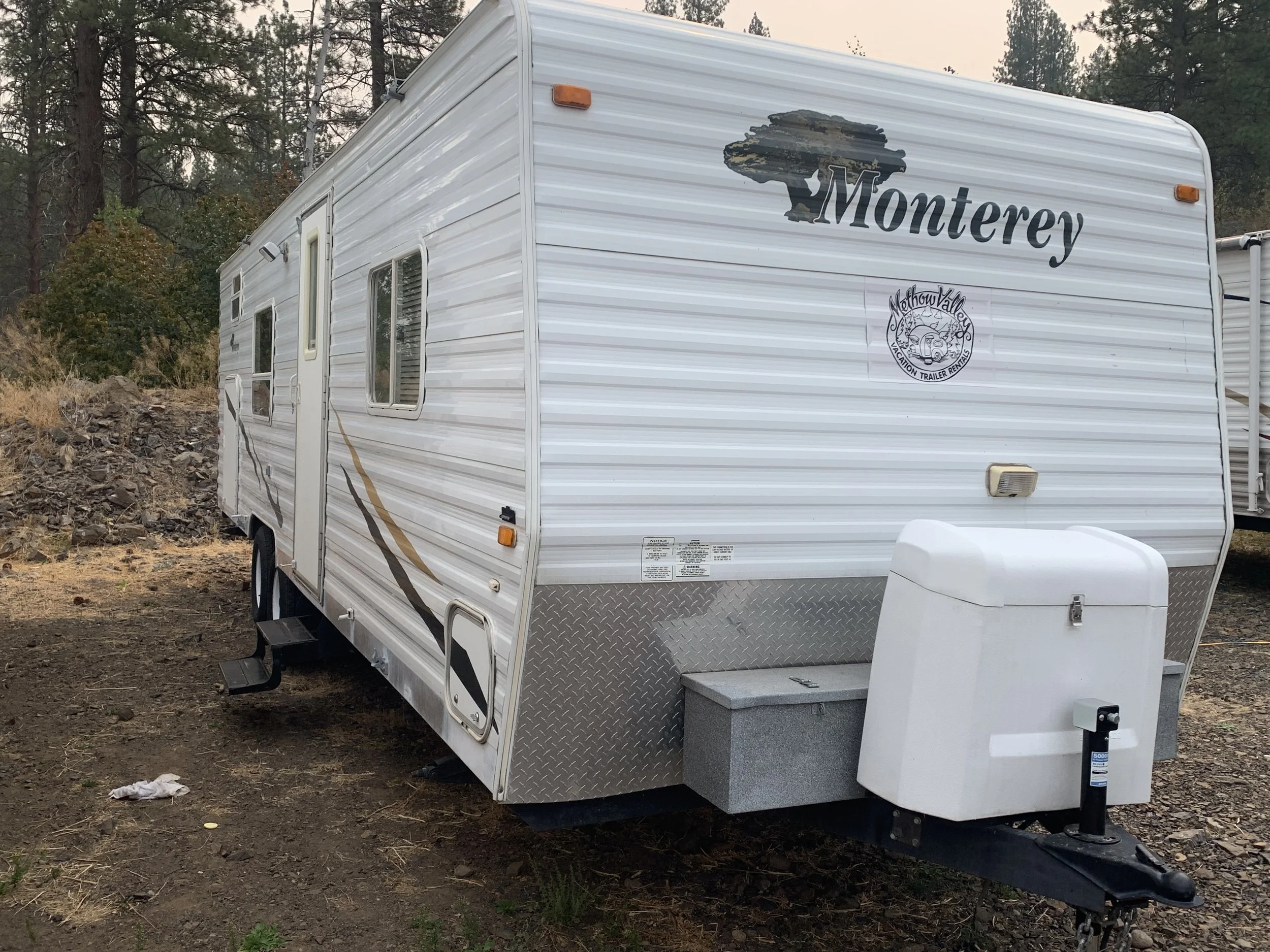 A white travel trailer with the word 'Monterey' and a tree logo on the front, parked on dirt ground with trees in the background.
