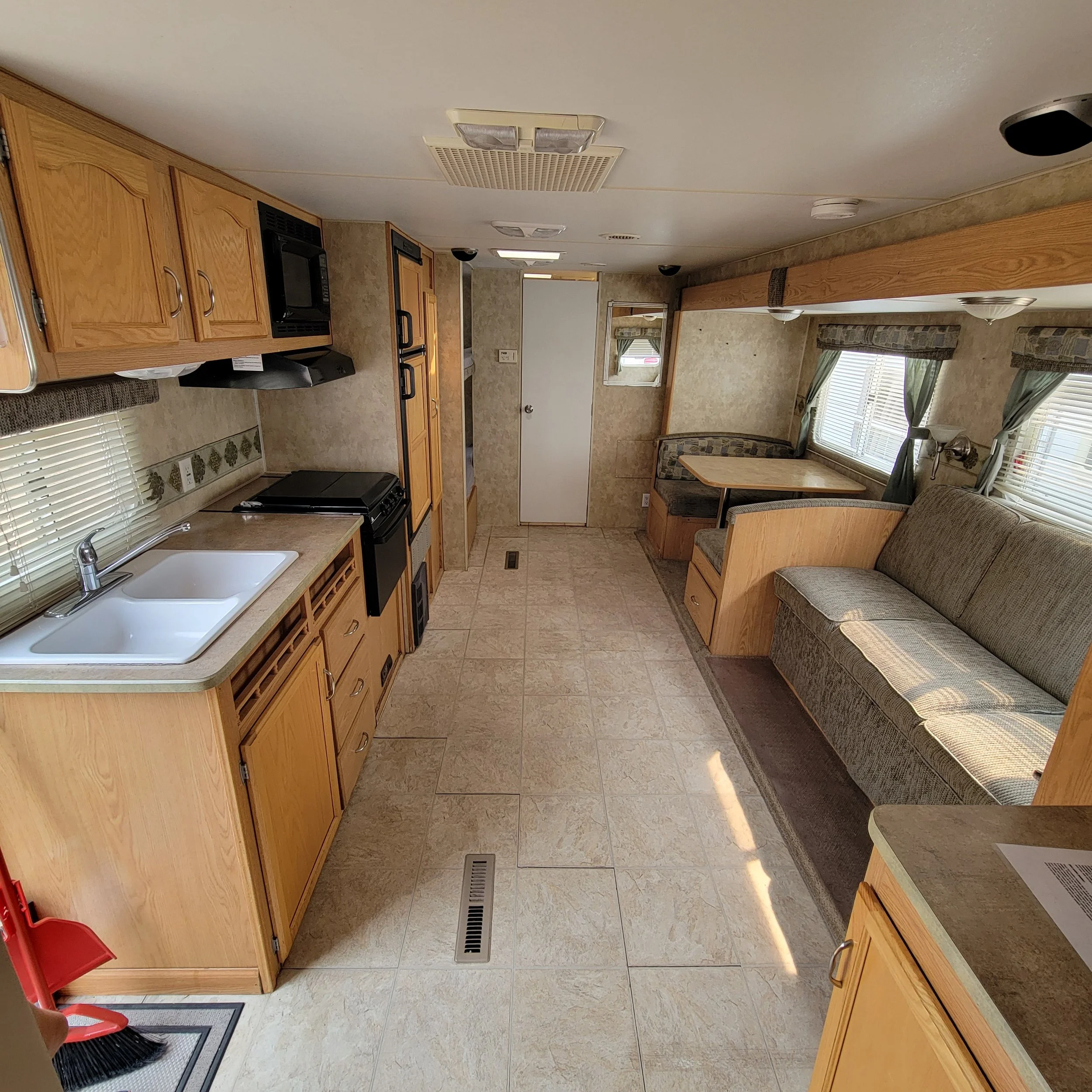 Interior of a small RV with a kitchen on the left, including a sink, stove, and cabinetry. On the right, there is a couch and a dining nook with windows and curtains. The floor has beige tiles, and the ceiling has vents and lighting fixtures.