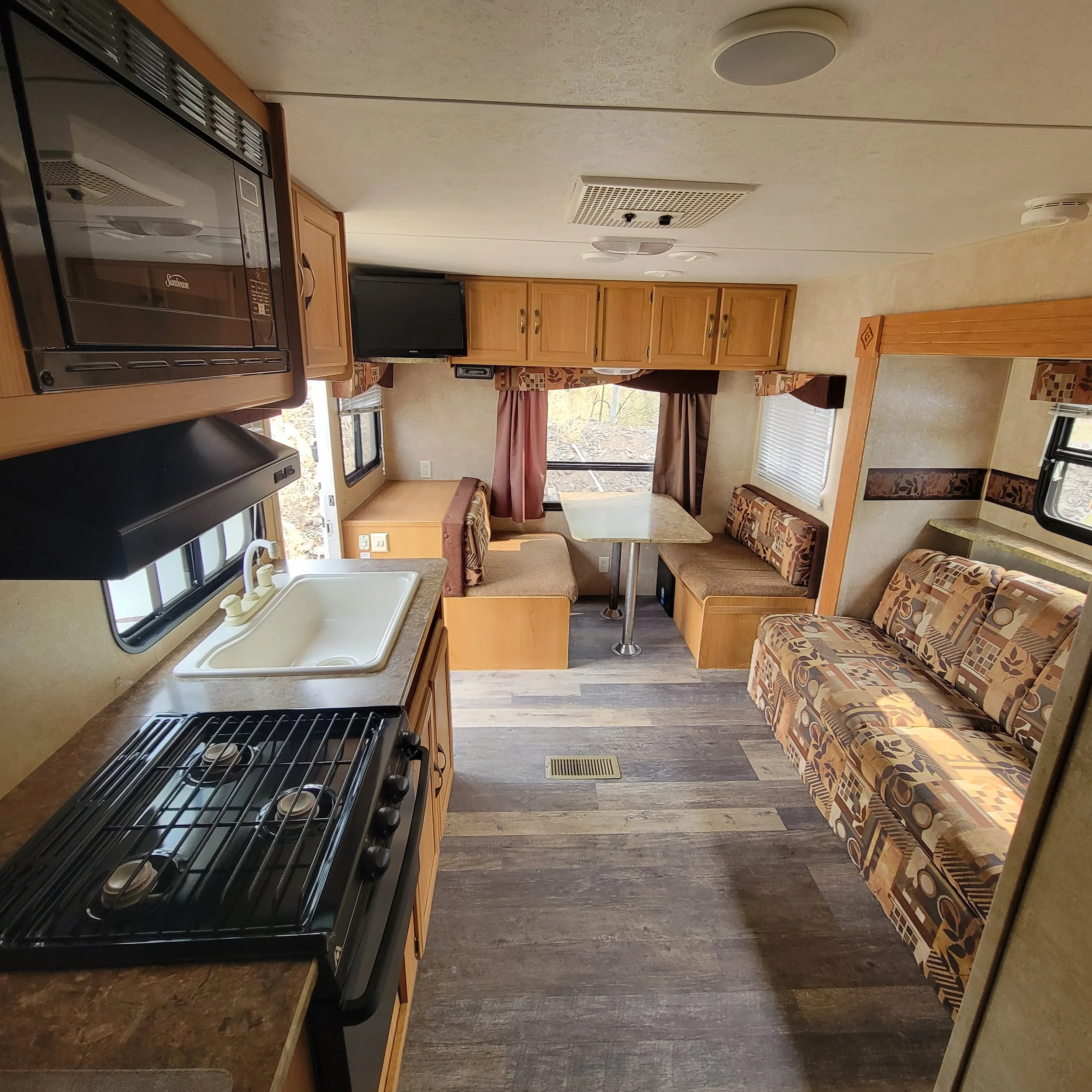Interior of a compact camper with a kitchen area: stove, sink, overhead microwave, dining booth, window with curtains, and a sofa with patterned upholstery.