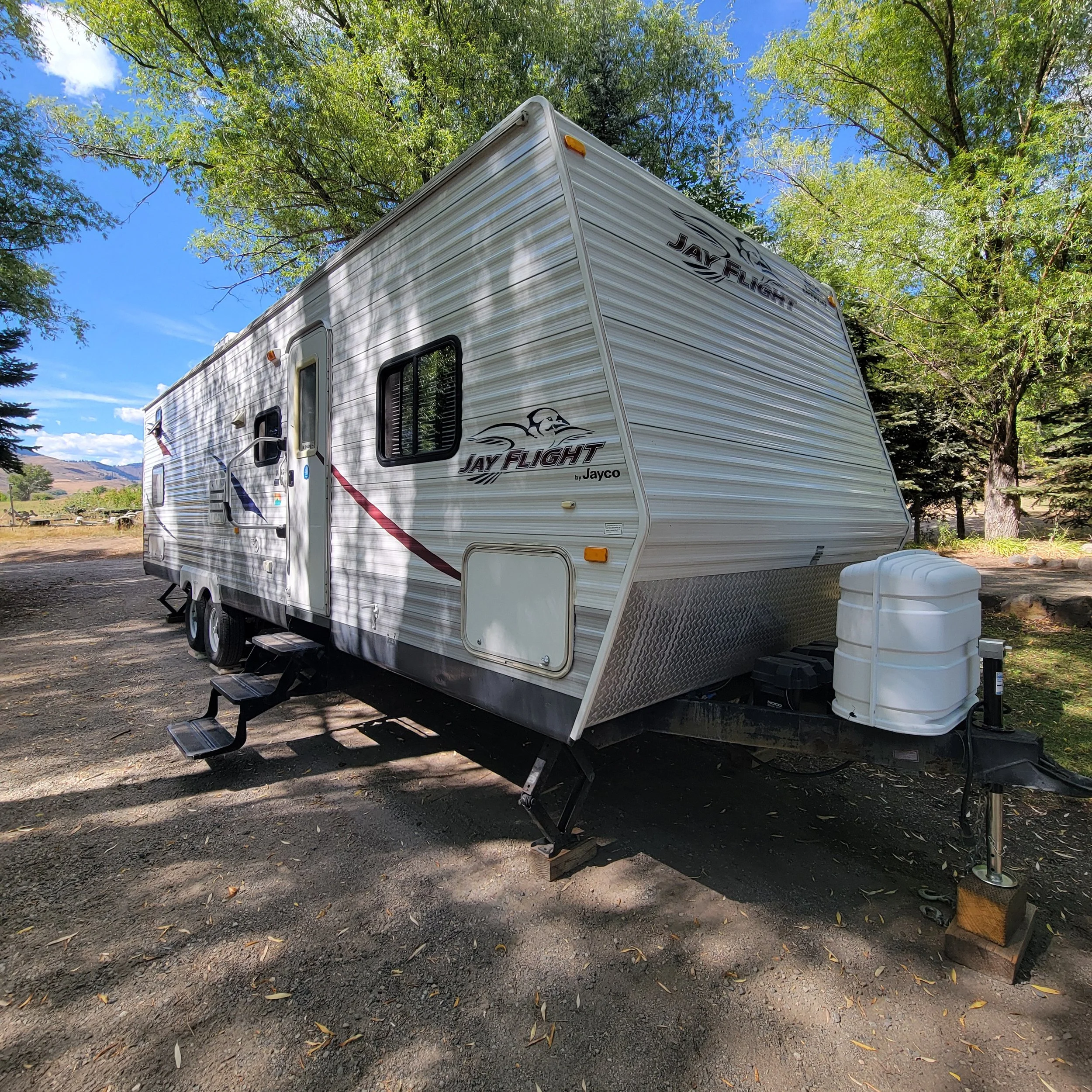 A white Jay Flight travel trailer parked on dirt ground outdoors, surrounded by green trees with a blue sky overhead.