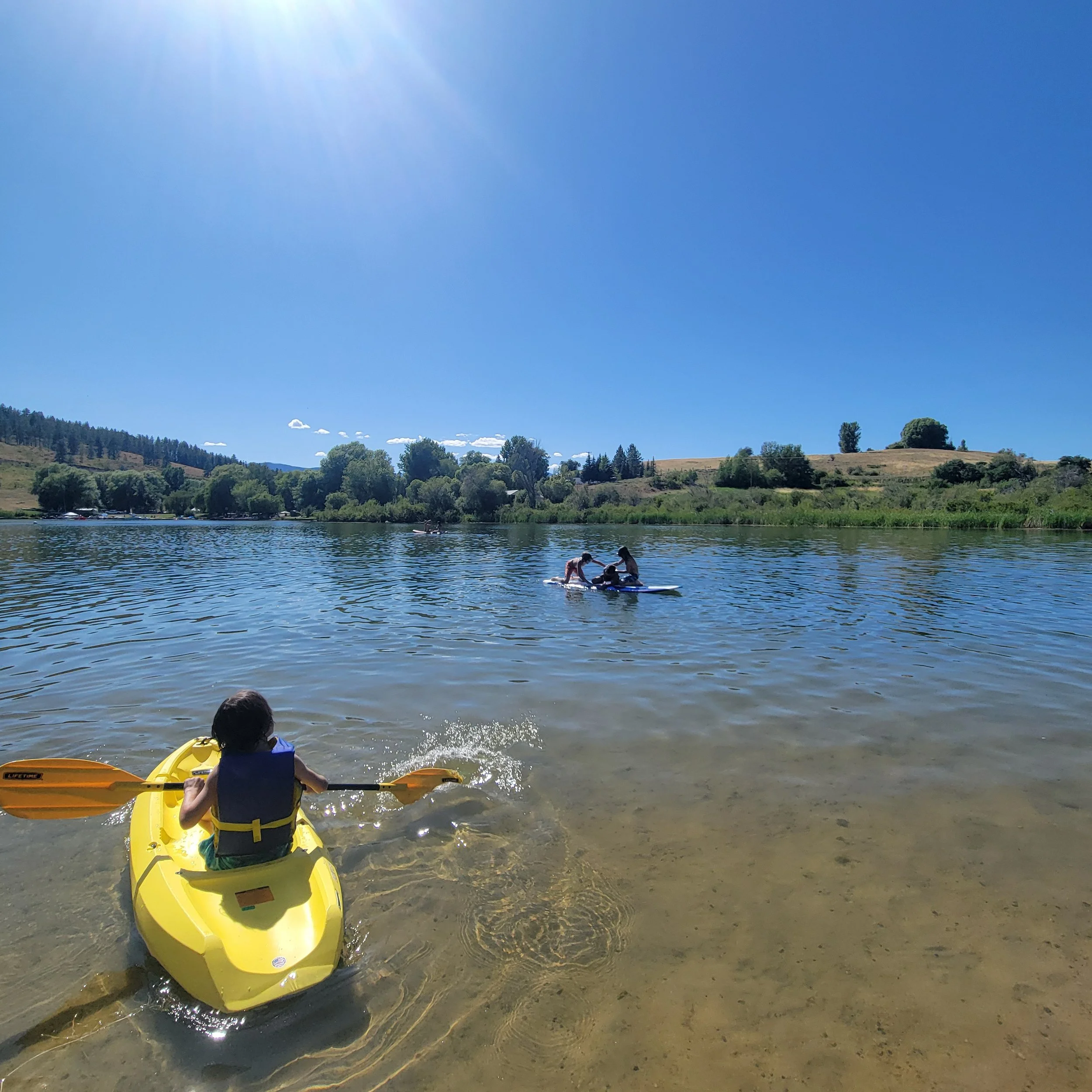 Child in a yellow kayak paddling in shallow water near the shoreline, with two people paddleboarding in the distance on a sunny day. Pearrygin Lake Winthrop WA. Fun outdoor sports