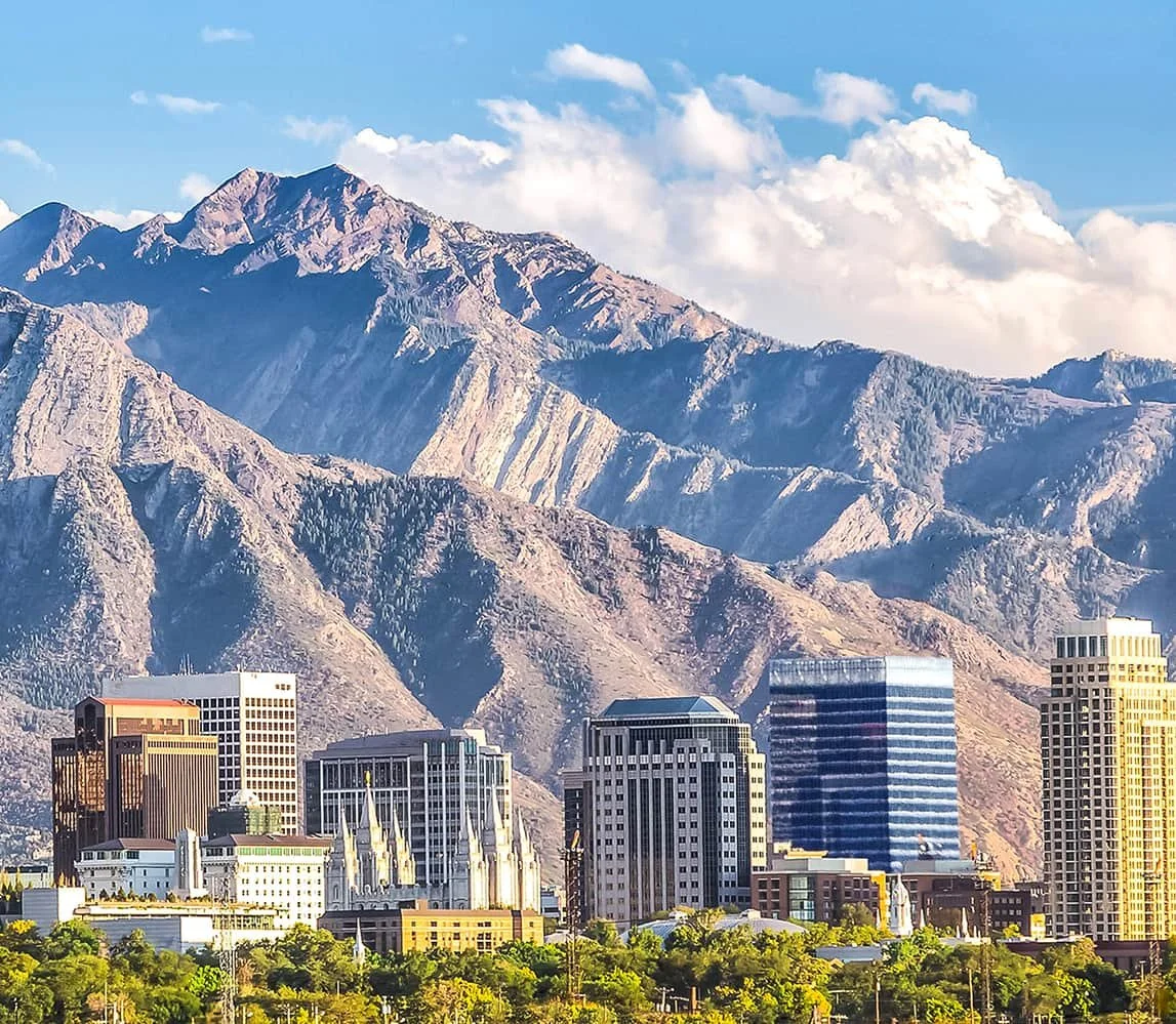 Skyline of Salt Lake City with modern buildings in front of the Wasatch Canyons under a partly cloudy sky.