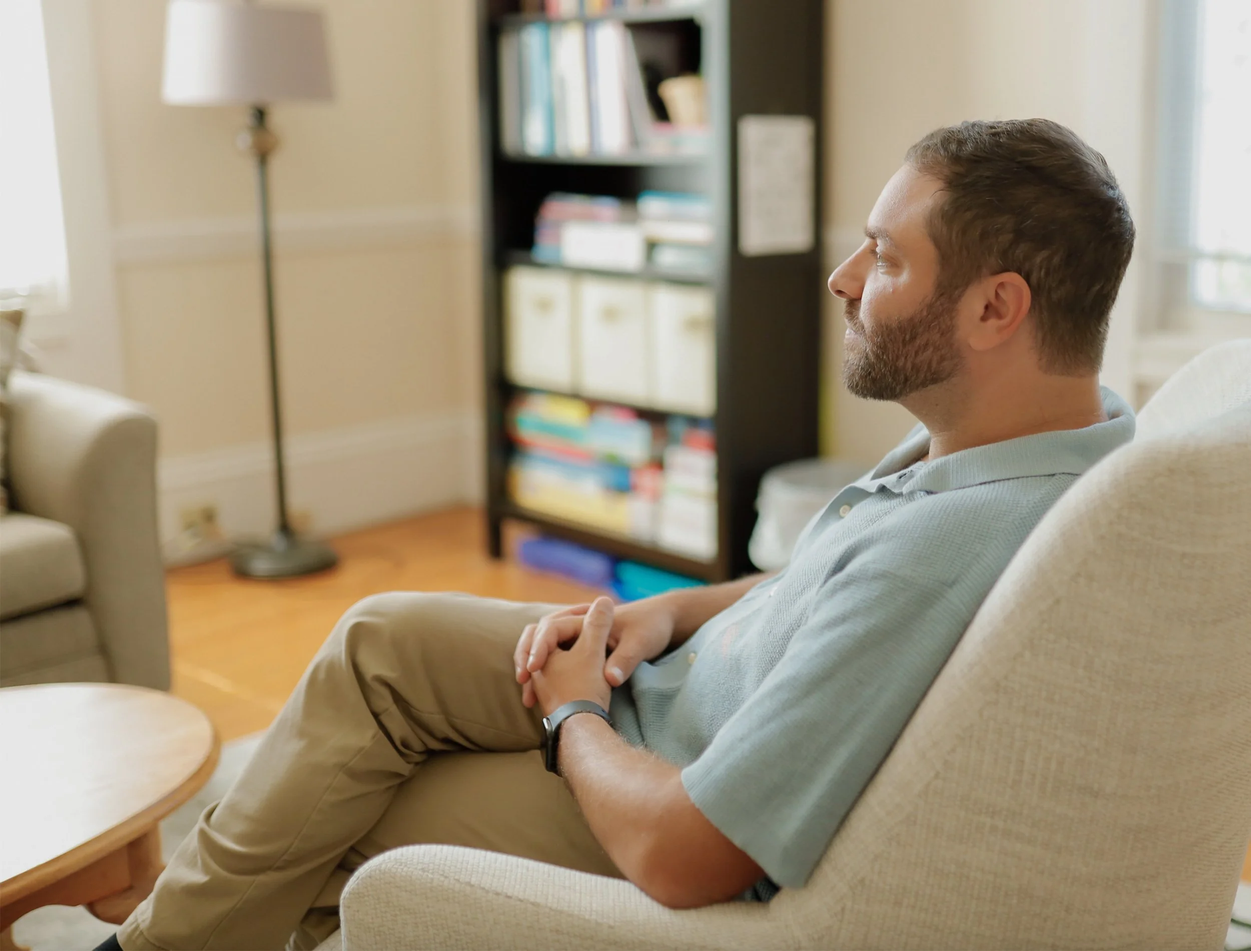 A man with short brown hair and a beard sitting on a beige armchair in a living room, with eyes closed, hands clasped, wearing a light green polo shirt and tan pants.