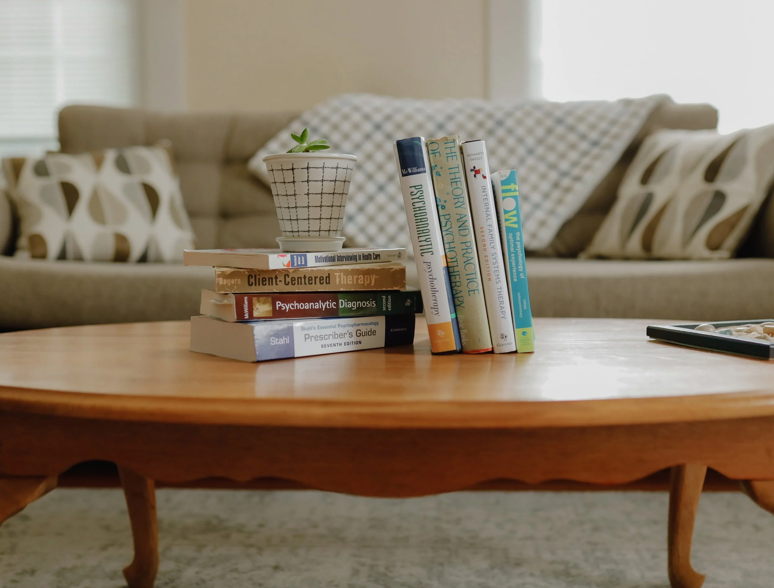 A wooden coffee table with books on psychology, therapy, and health, a small potted succulent, and a tray of pills, in front of a beige couch with patterned pillows in a living room.