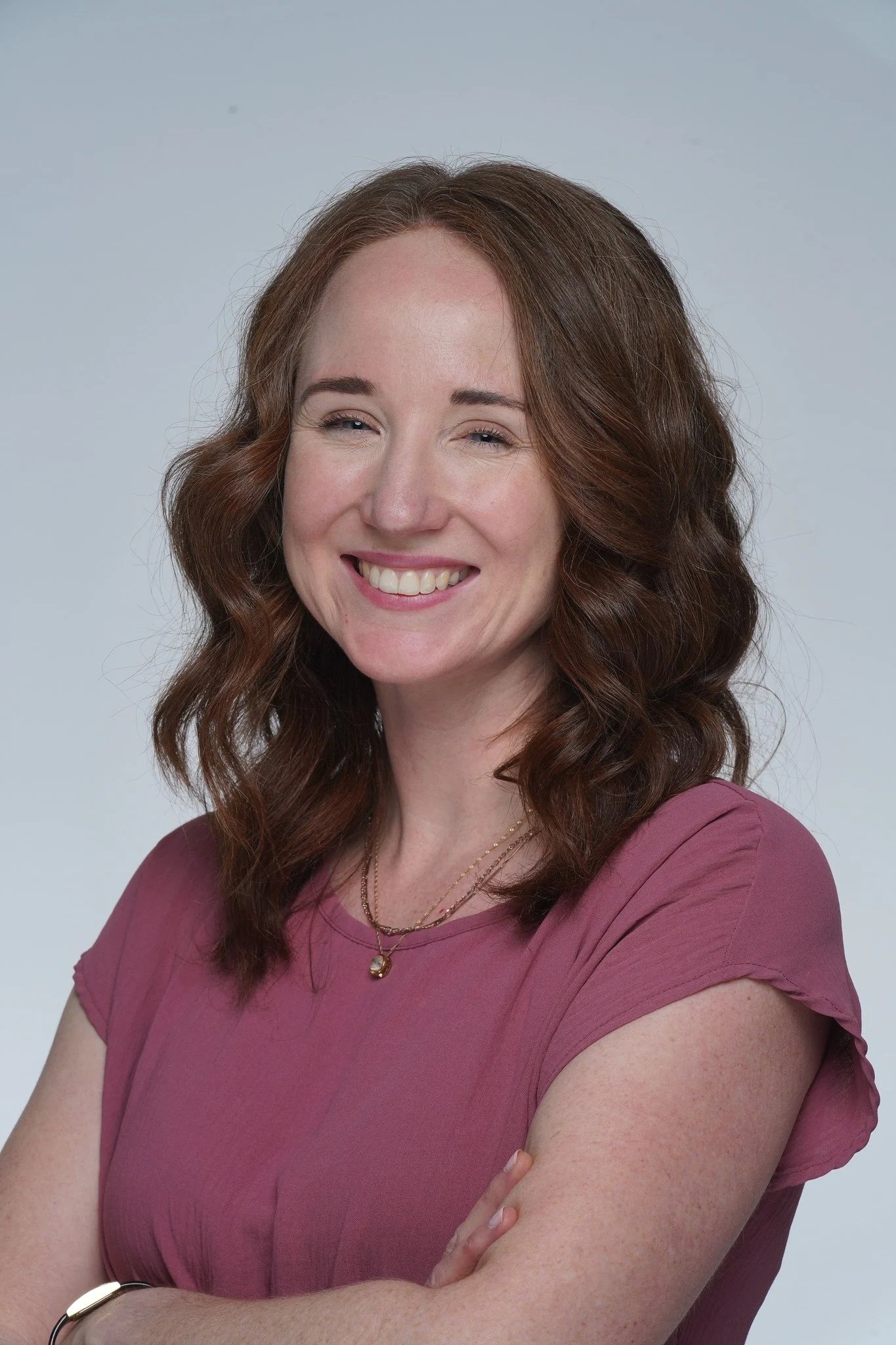 A woman with shoulder-length wavy red hair, smiling, wearing a pinkish-mauve top, gold necklace, and a bracelet, posing with crossed arms against a plain white background.