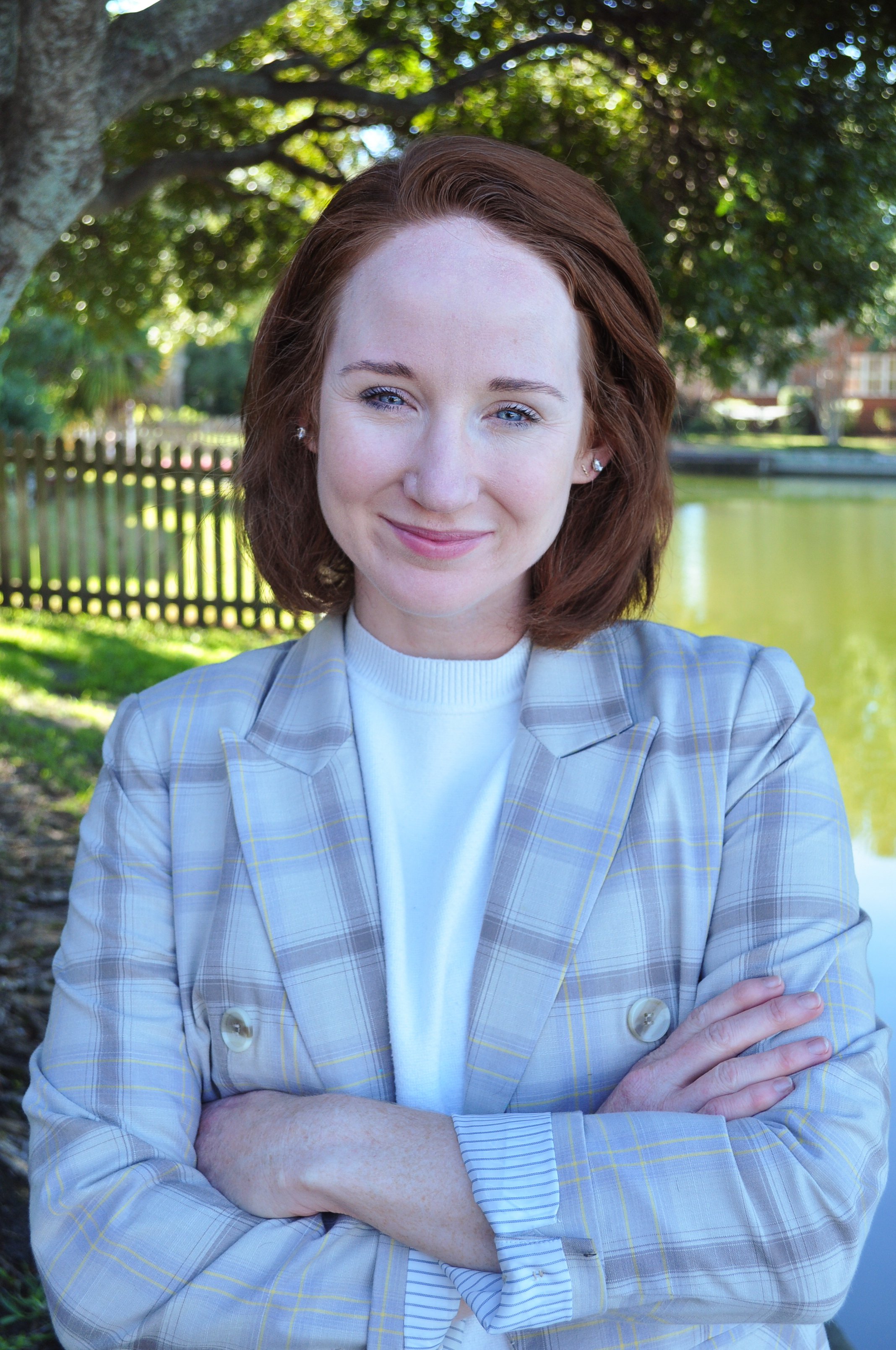Lisa Cherfane, Marketing Strategist, wearing a light gray checkered blazer and crossing her arms. She is smiling and the scene includes trees and a fence in the background.