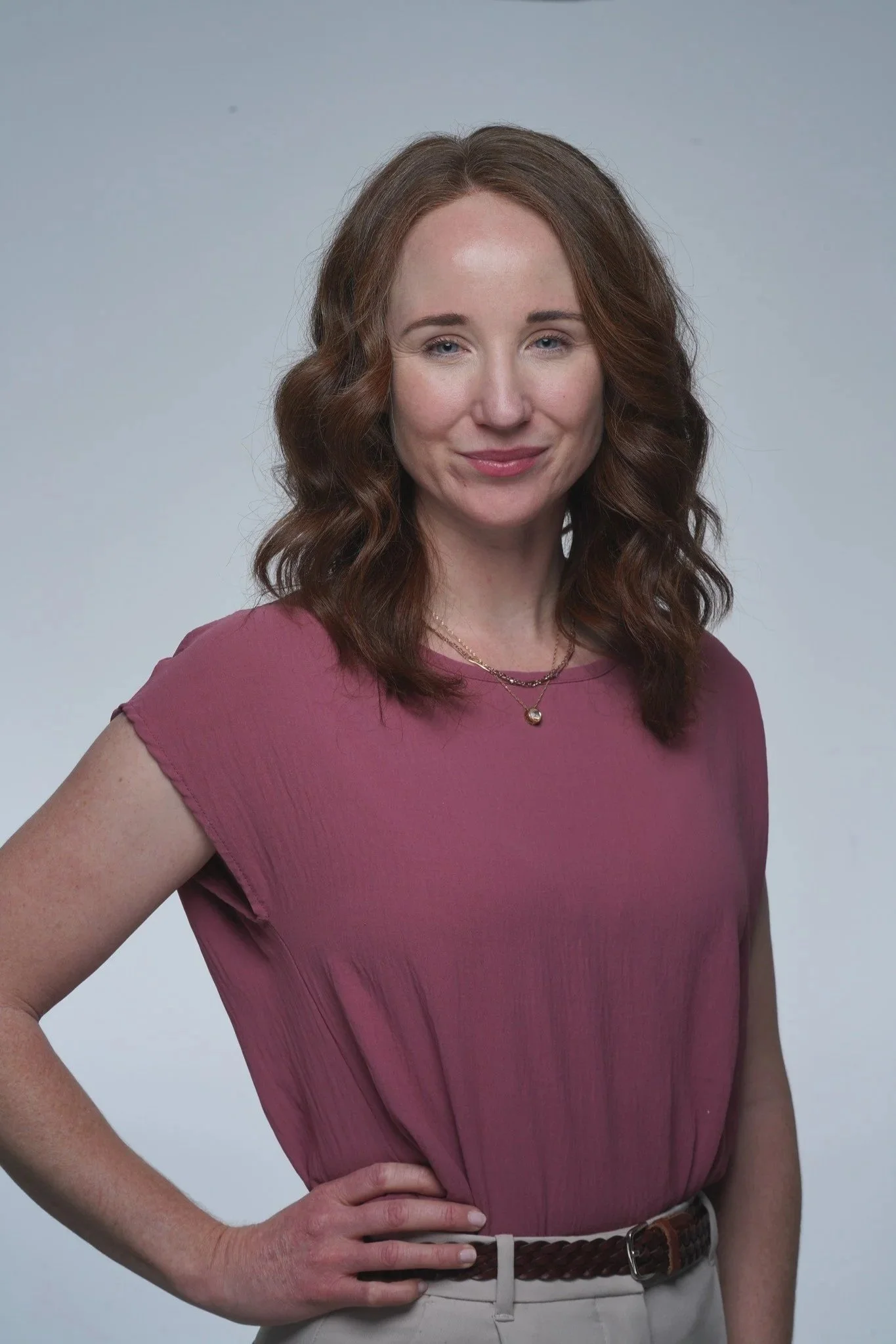 Lisa Cherfane, Branding Strategist, wearing a pink blouse, beige pants, and a brown braided belt, stands against a light grey background with a confident expression.