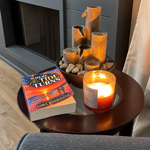 A copy of Where the Tide Turns beside a glowing candle and a fireplace.