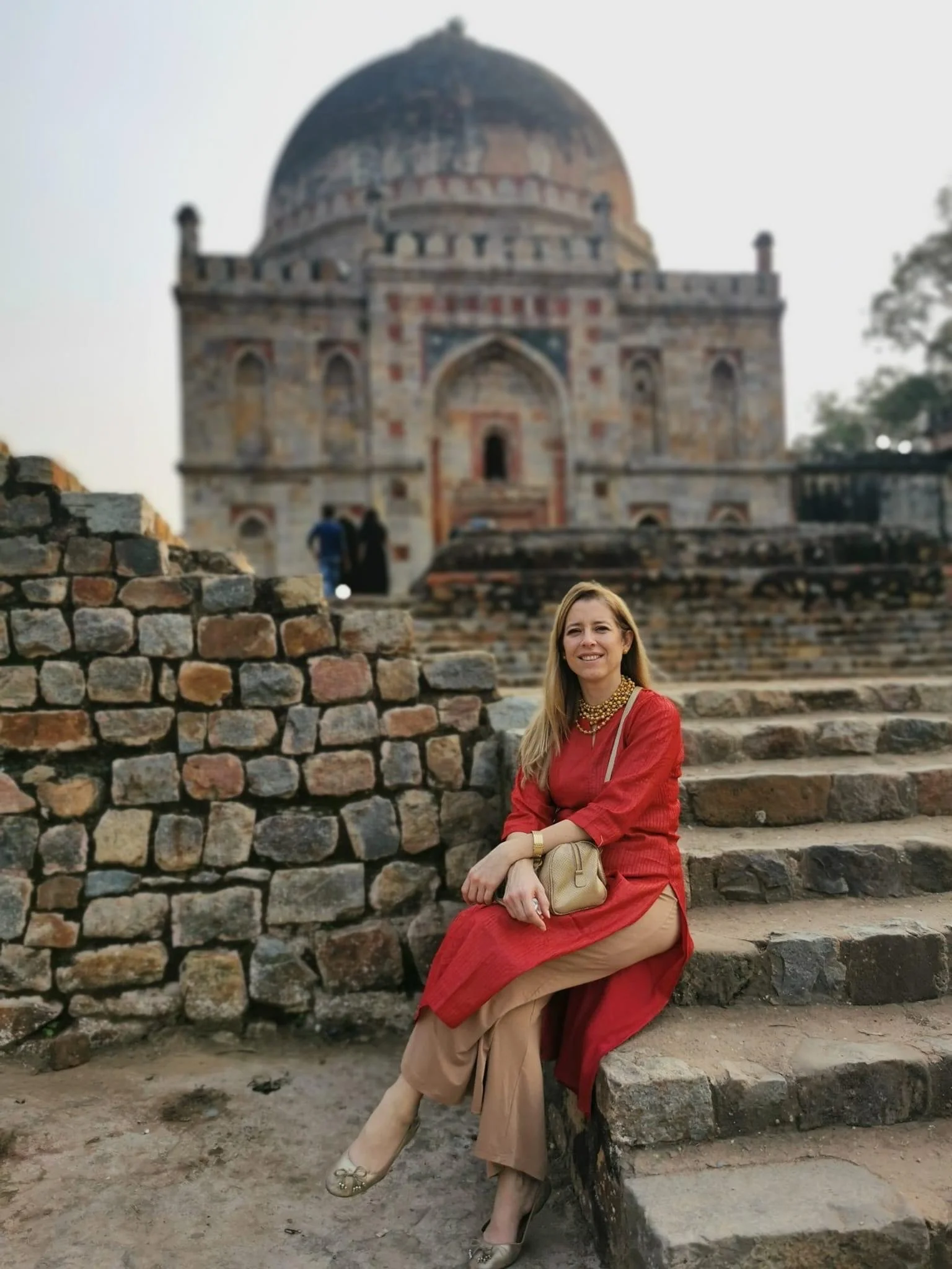 A woman in a red dress sitting on stone steps in front of an ancient building with a large dome, likely at a historical site in India.