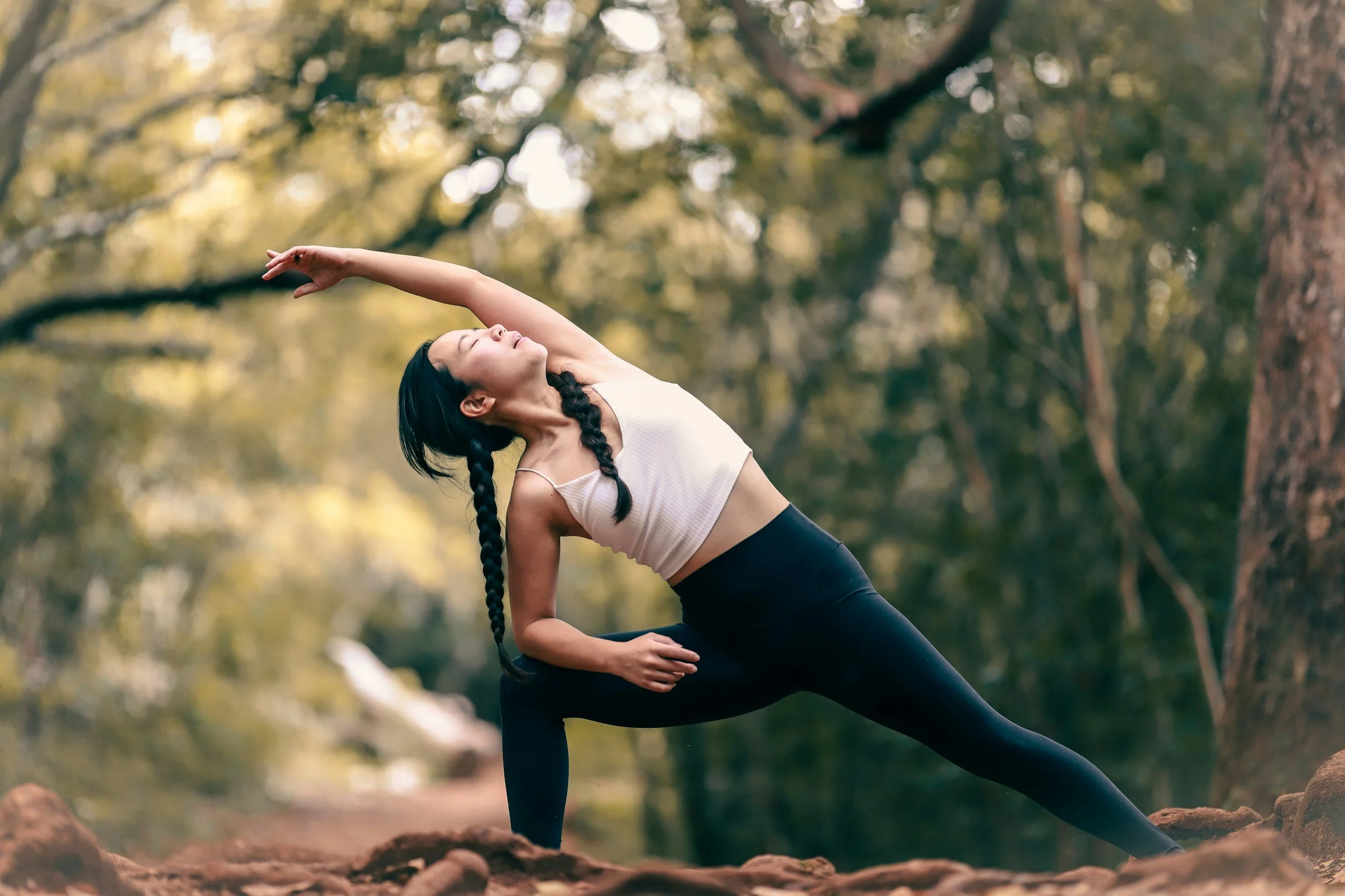 A woman with long braided hair practicing yoga outdoors on a rocky terrain in a forest, performing a side stretch pose.