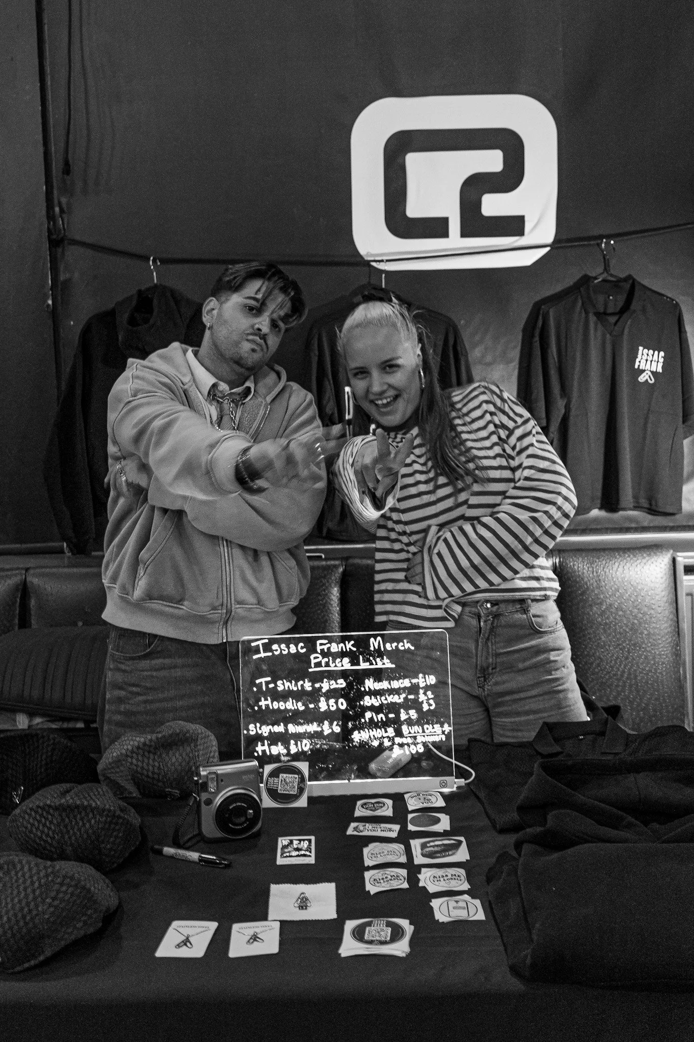 Two people posing behind a table selling merchandise, including stickers, patches, and pins, with a sign listing prices. Behind them, clothes are hanging with one shirt labeled 'Isaac Frank Merch.'