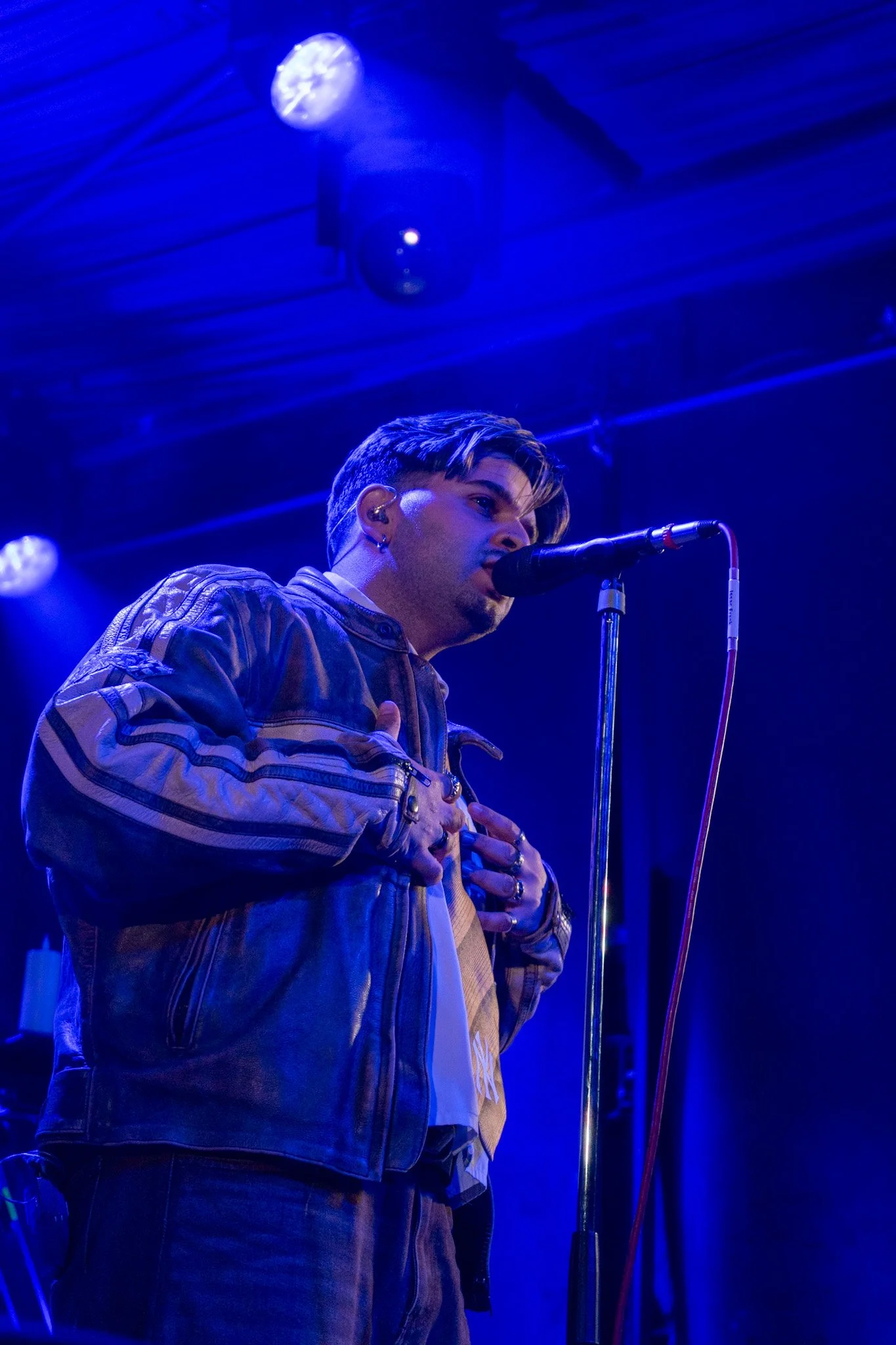 A male singer performing on stage under blue lights, wearing a brown jacket, holding his hands over his chest, with a microphone in front of him.