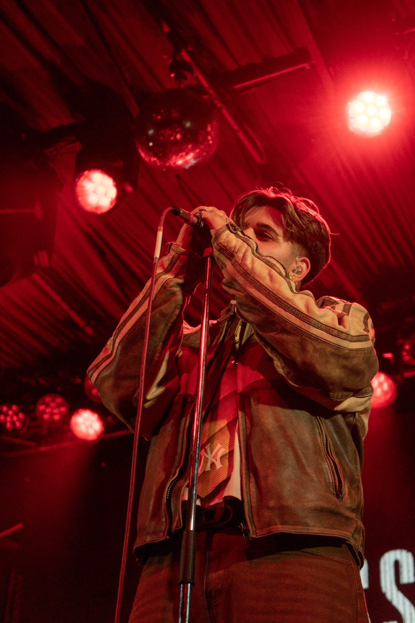A young man singing into a microphone on stage, red lighting and a disco ball overhead.