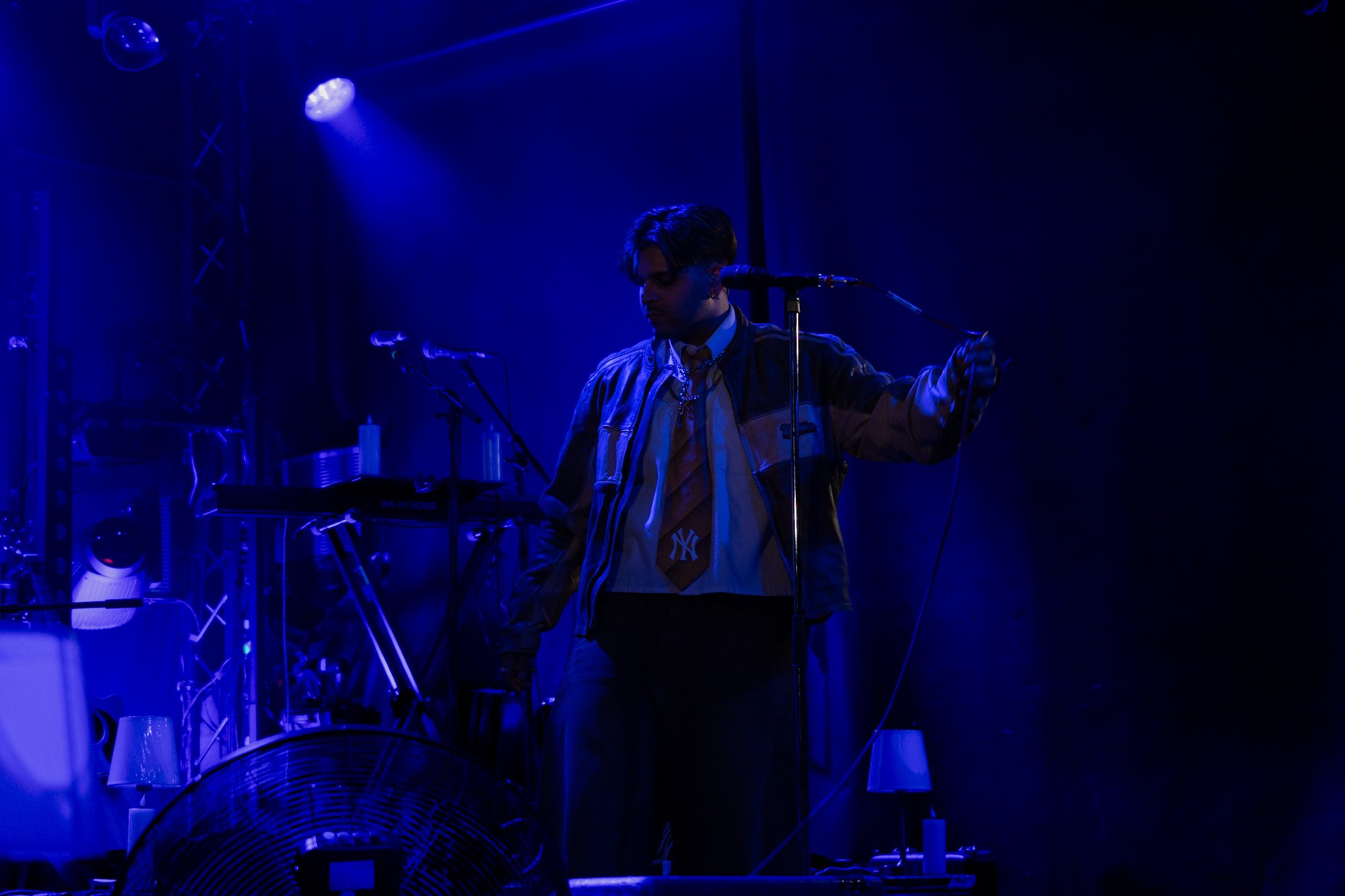 A person with dark hair wearing a jacket and tie, standing in front of microphones on a dark stage illuminated by blue lighting, with electronic equipment and a fan visible in the background.