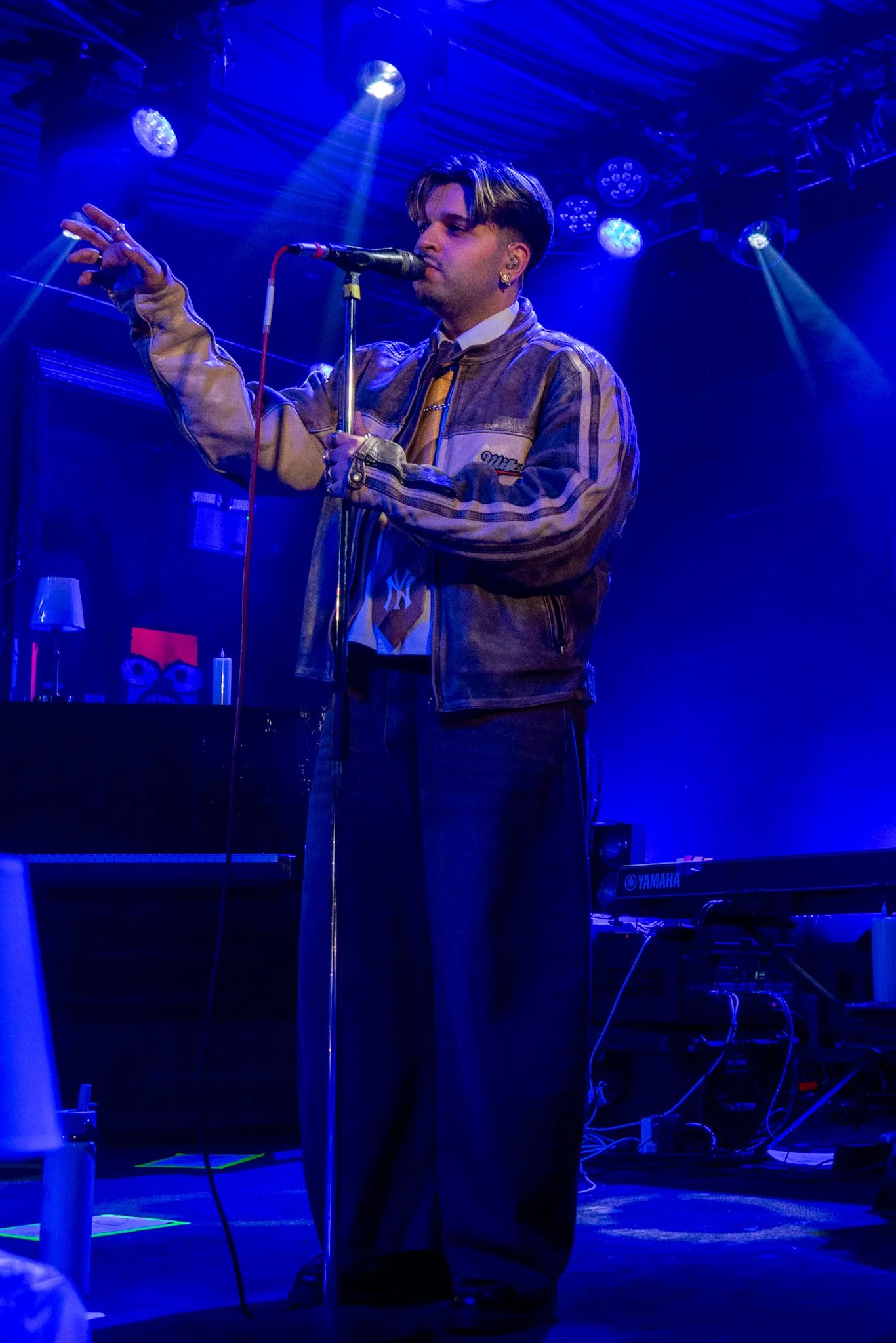 A young man singing into a microphone on stage with blue lighting, wearing a jacket, earrings, and a shirt with a tie.