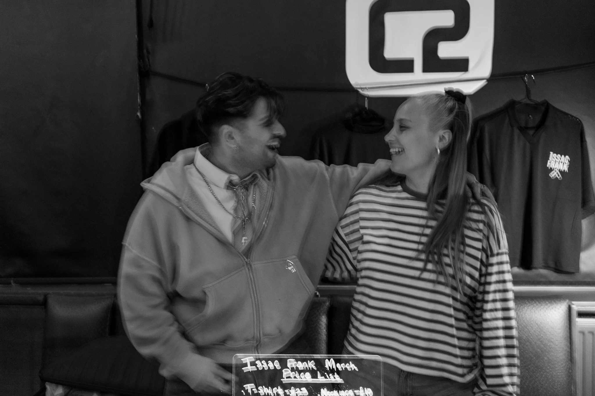 Two people, a man and a woman, smiling and looking at each other, standing behind a glass display case with a sign that says 'Icee, Frank Merch, Price List', in a store or market.