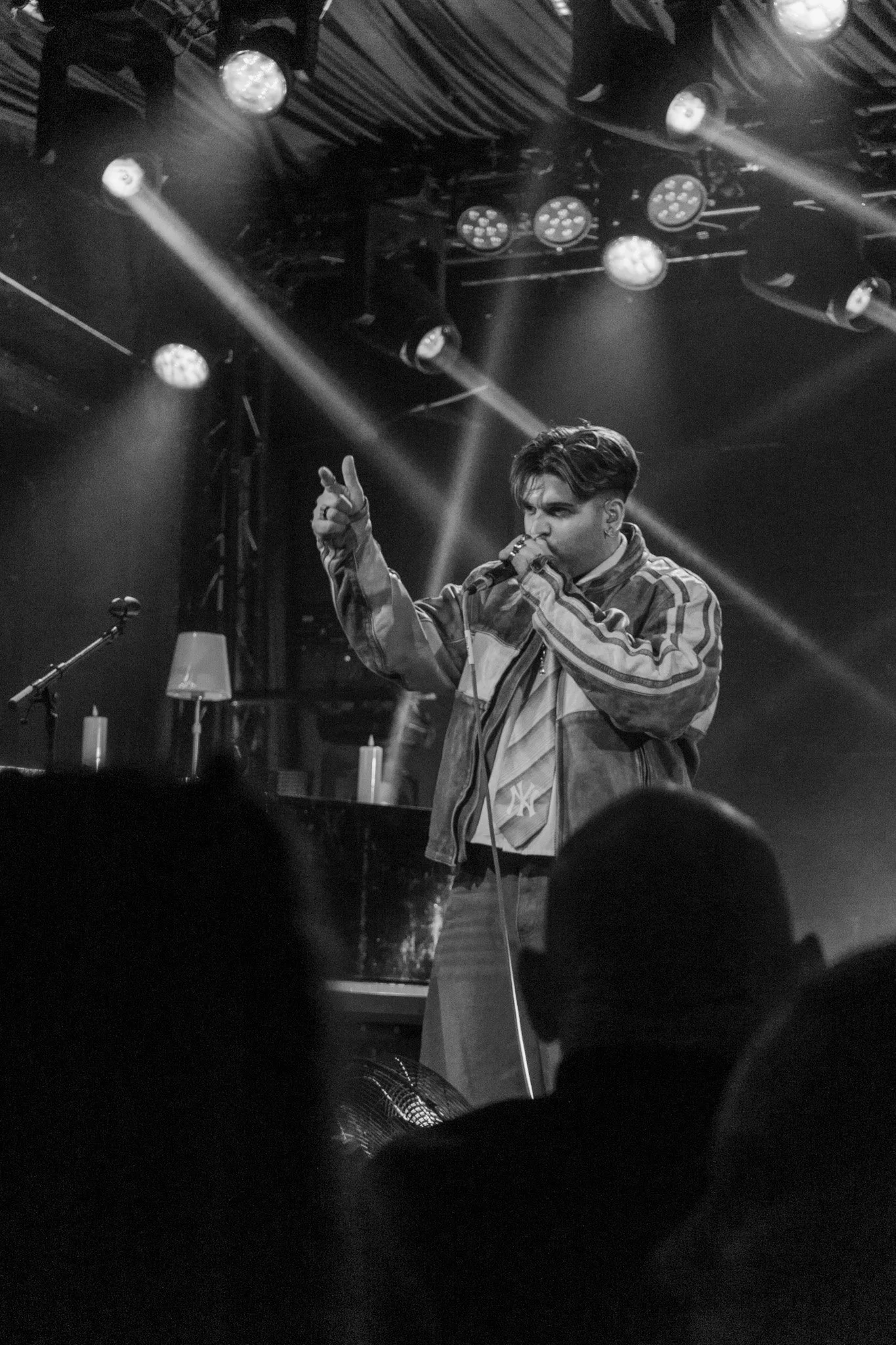 A male singer performing on stage with a microphone, wearing a striped jacket and plaid pants, under bright stage lights in a dark venue.