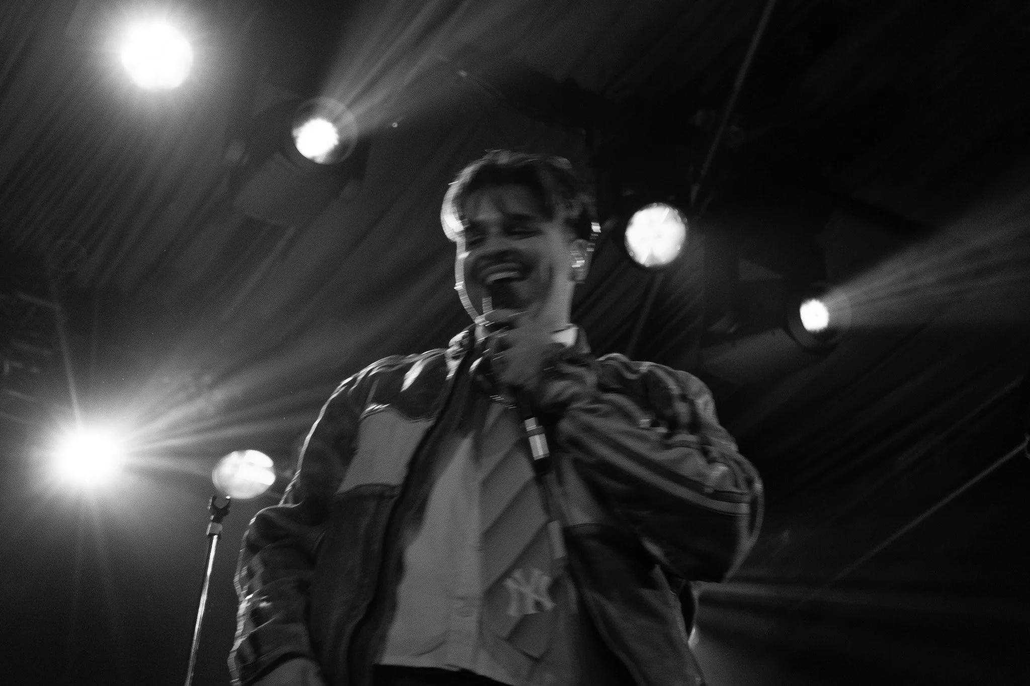Black and white photo of a young man smiling and singing into a microphone on stage, with bright stage lights overhead.