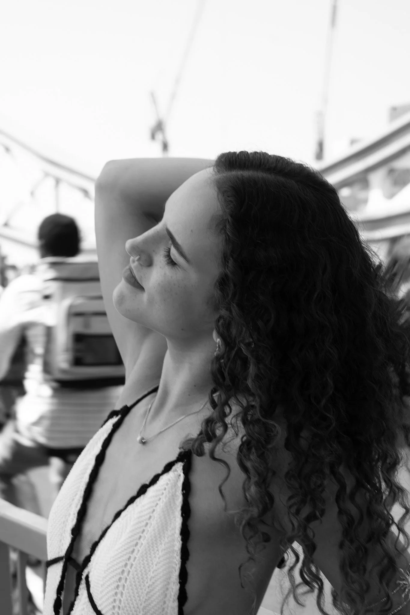 A woman with curly hair smiling with her eyes closed, wearing a white sleeveless top and a necklace, in an outdoor setting with blurred people and structures in the background.