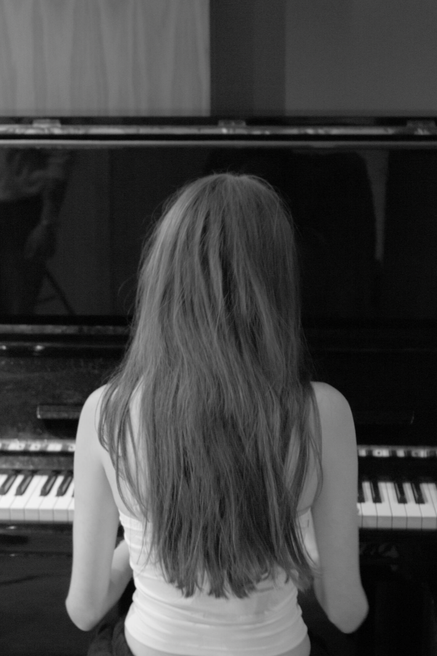 A woman with long hair sitting and playing piano, seen from behind, in black and white.
