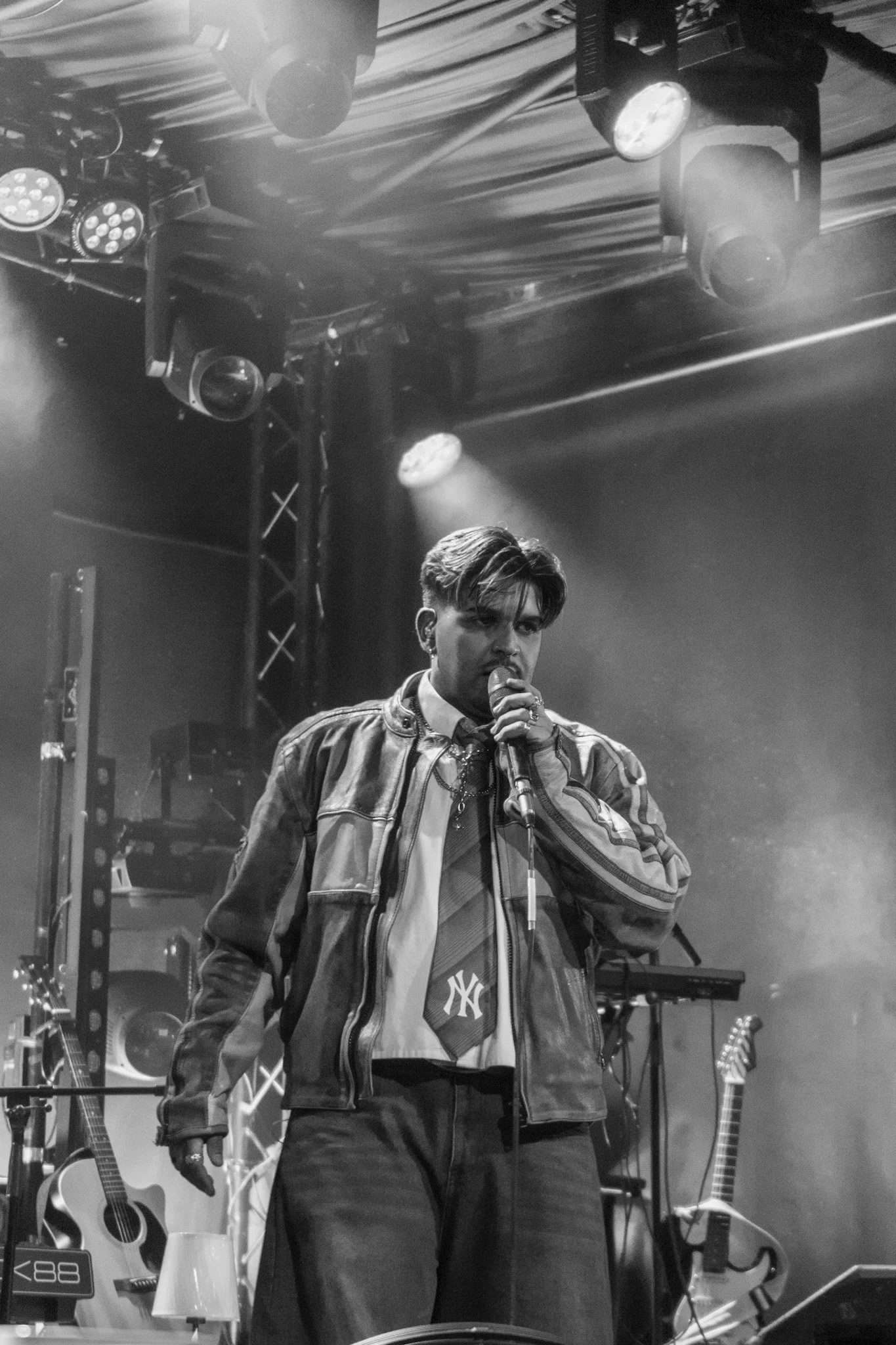 A male singer with styled hair and earrings performing on stage, holding a microphone, with musical instruments and stage lighting in the background.