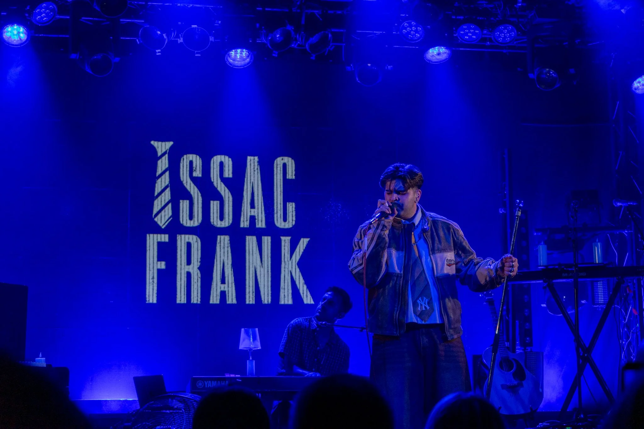 A male performer singing into a microphone on stage with blue lighting, a keyboard, and a guitar, in front of a backdrop that says 'ISSAC FRANK'.
