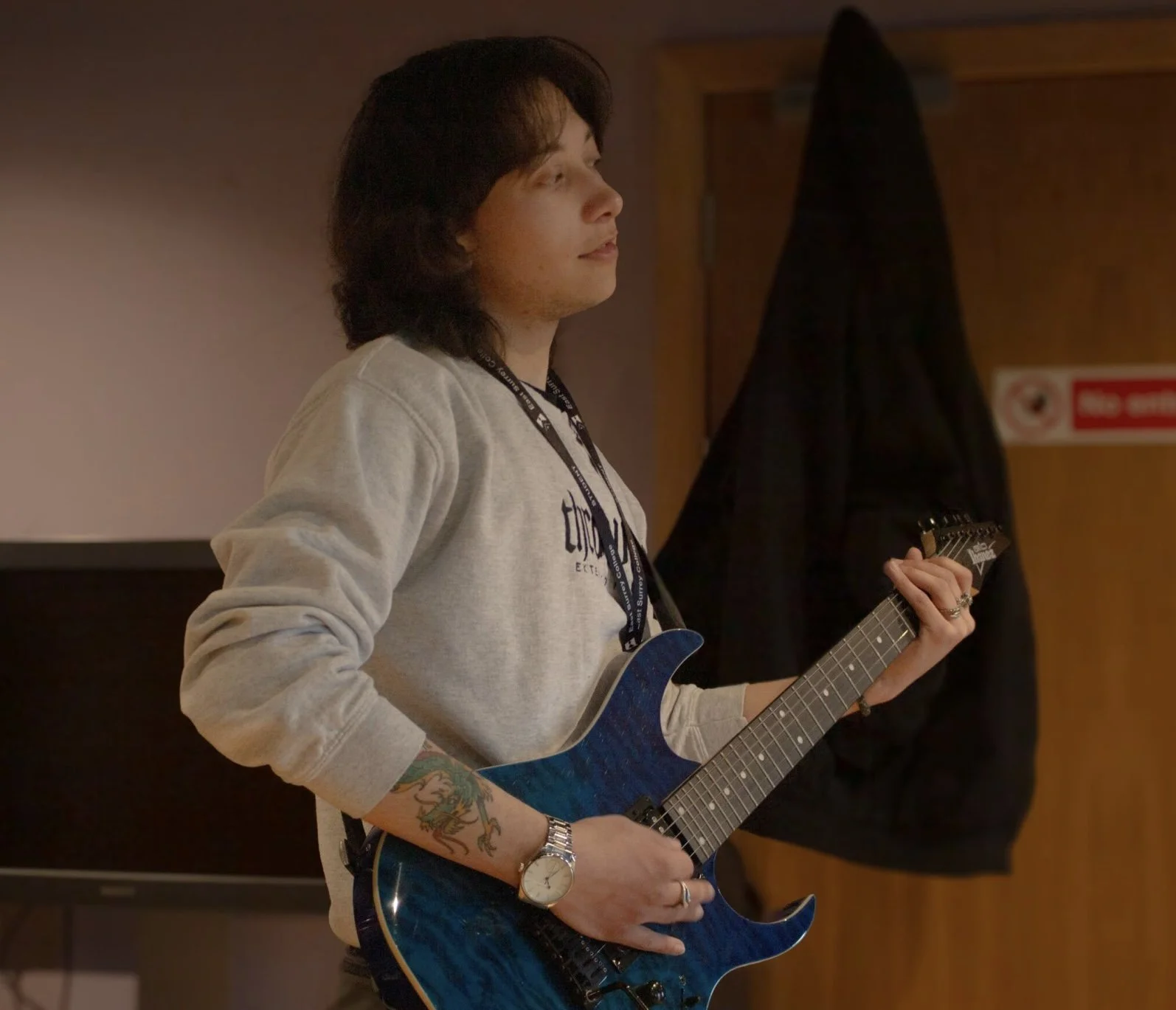 A young man with dark hair playing a blue electric guitar, wearing a gray sweatshirt, standing indoors with a wooden door and black jacket in the background.