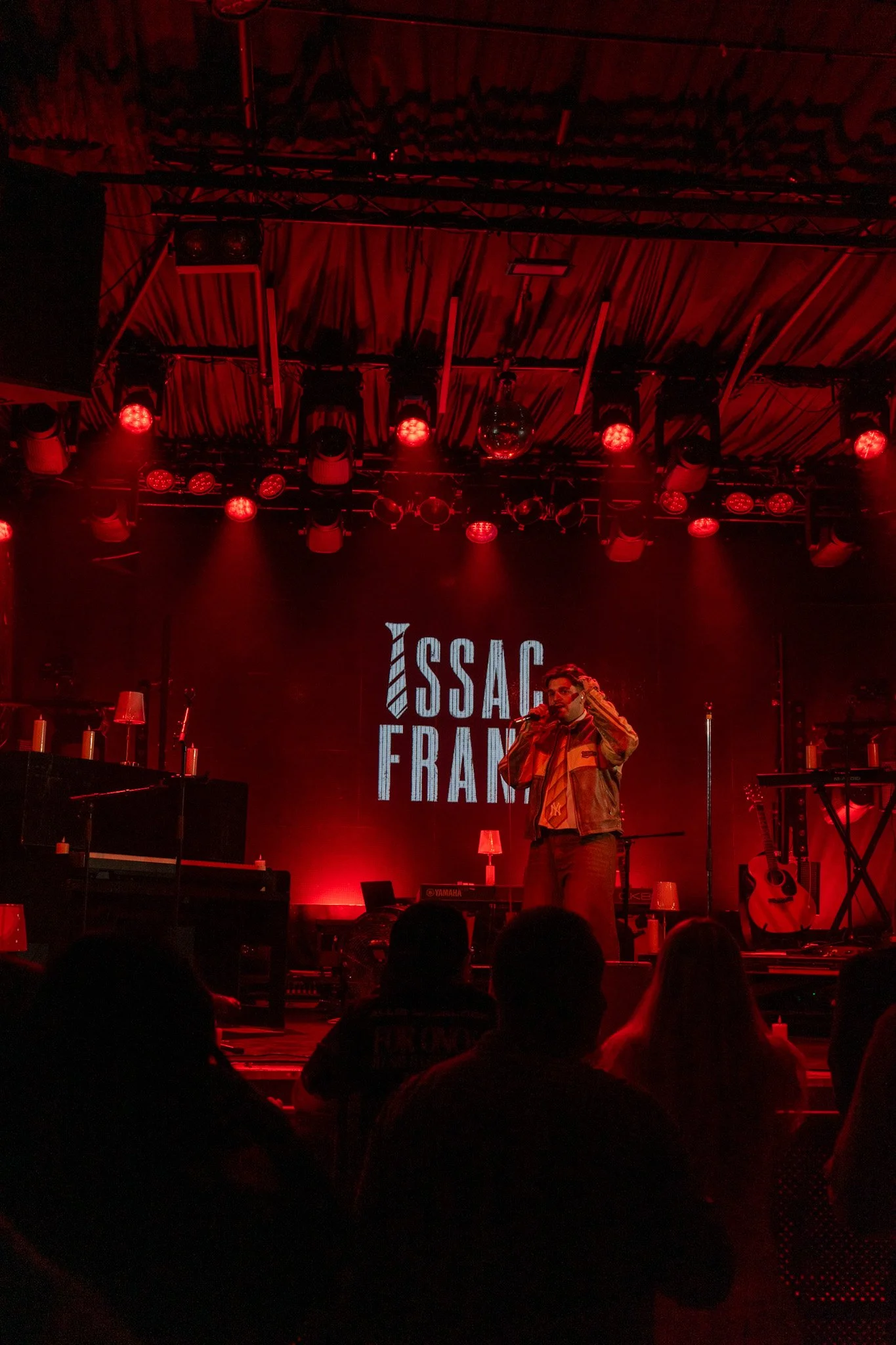 A stage with red lighting, musical instruments, and a large screen displaying the words 'SSAC FRAM'. A performer is standing in front of the screen, speaking or singing into a microphone. Audience members are visible in silhouette in the foreground.