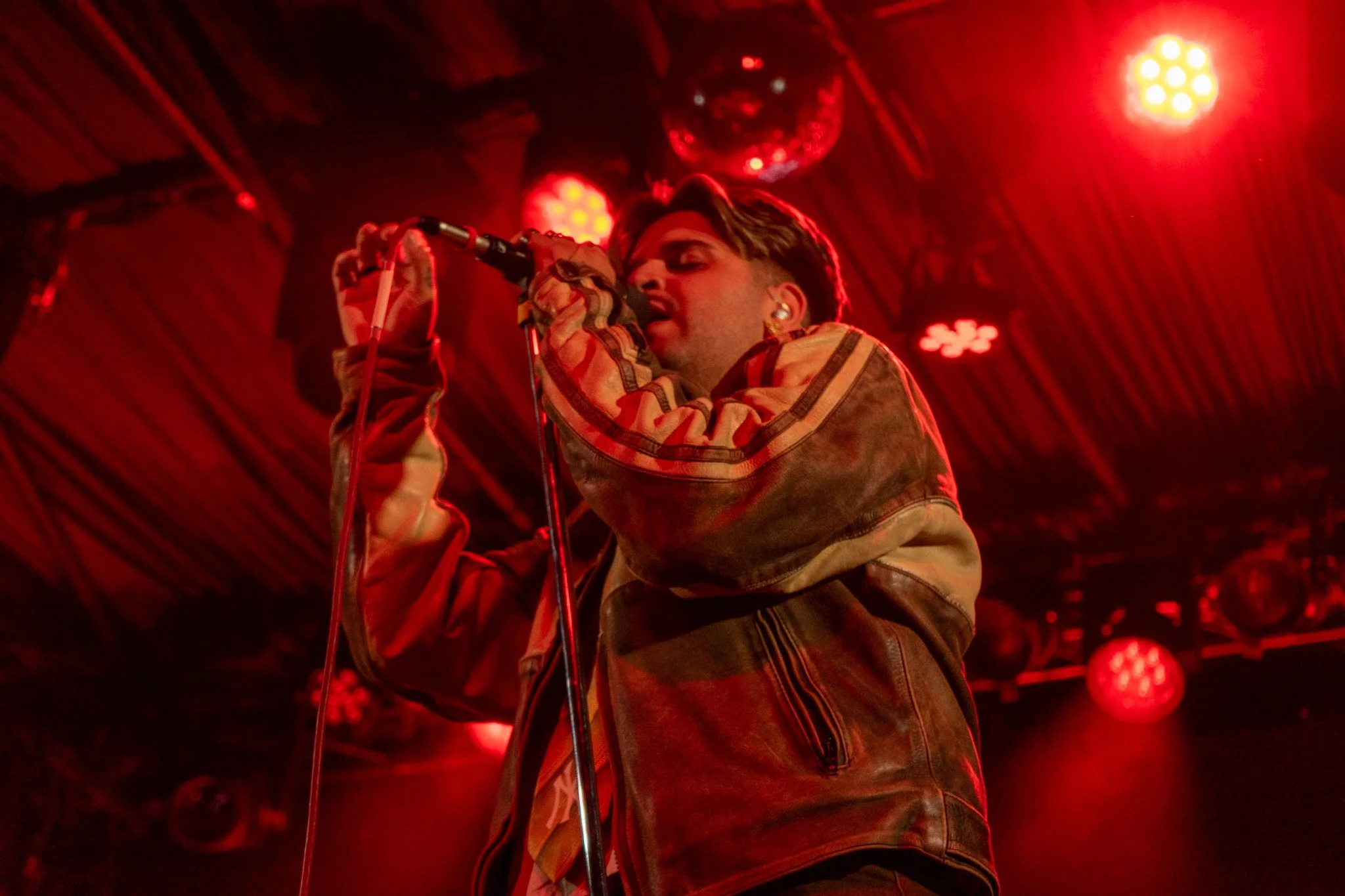 A person singing into a microphone on stage, illuminated by red stage lights.
