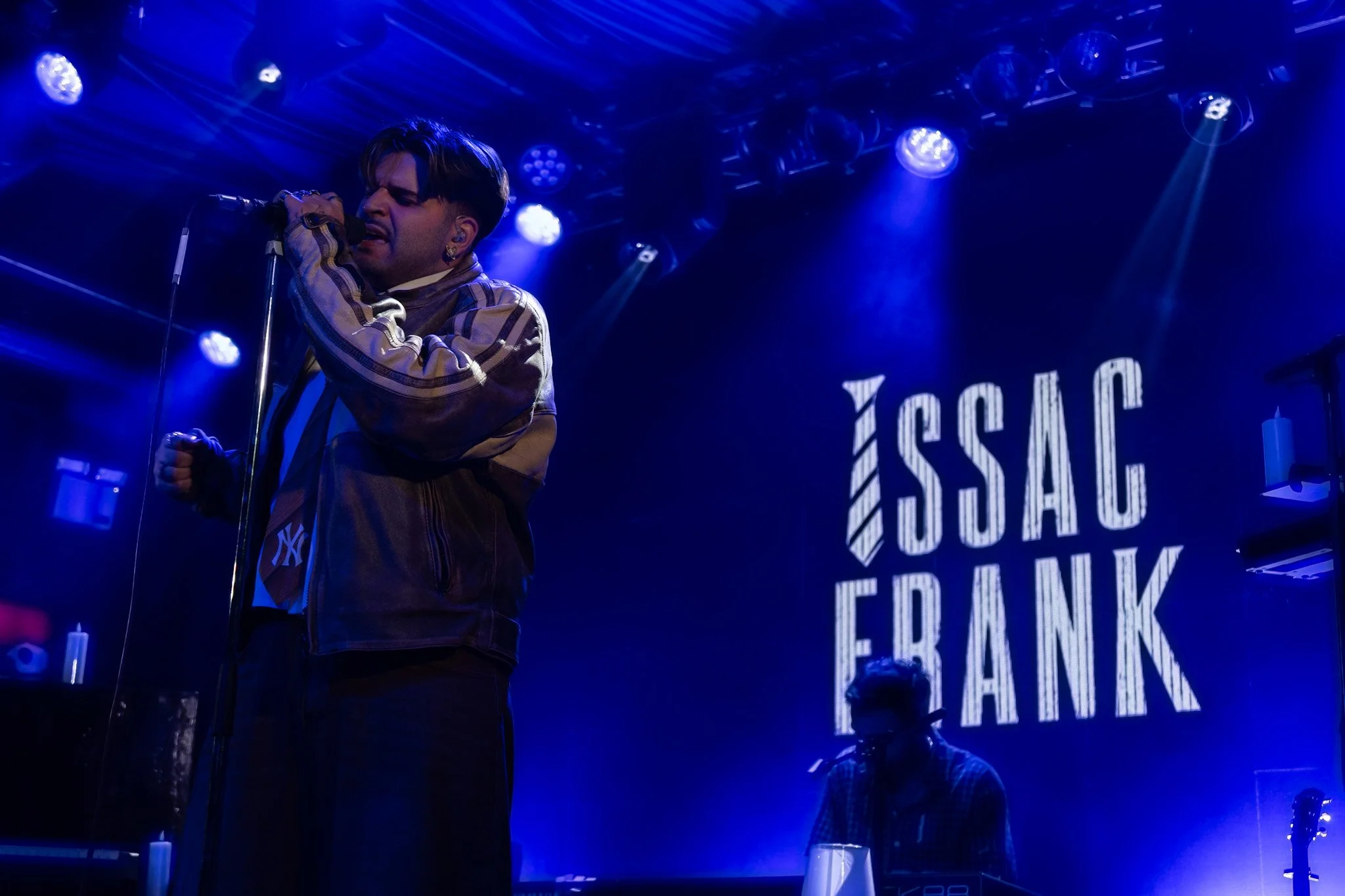 A male singer performing on stage with blue lighting, holding a microphone, with the words 'ESCAPE' and other text projected in the background.
