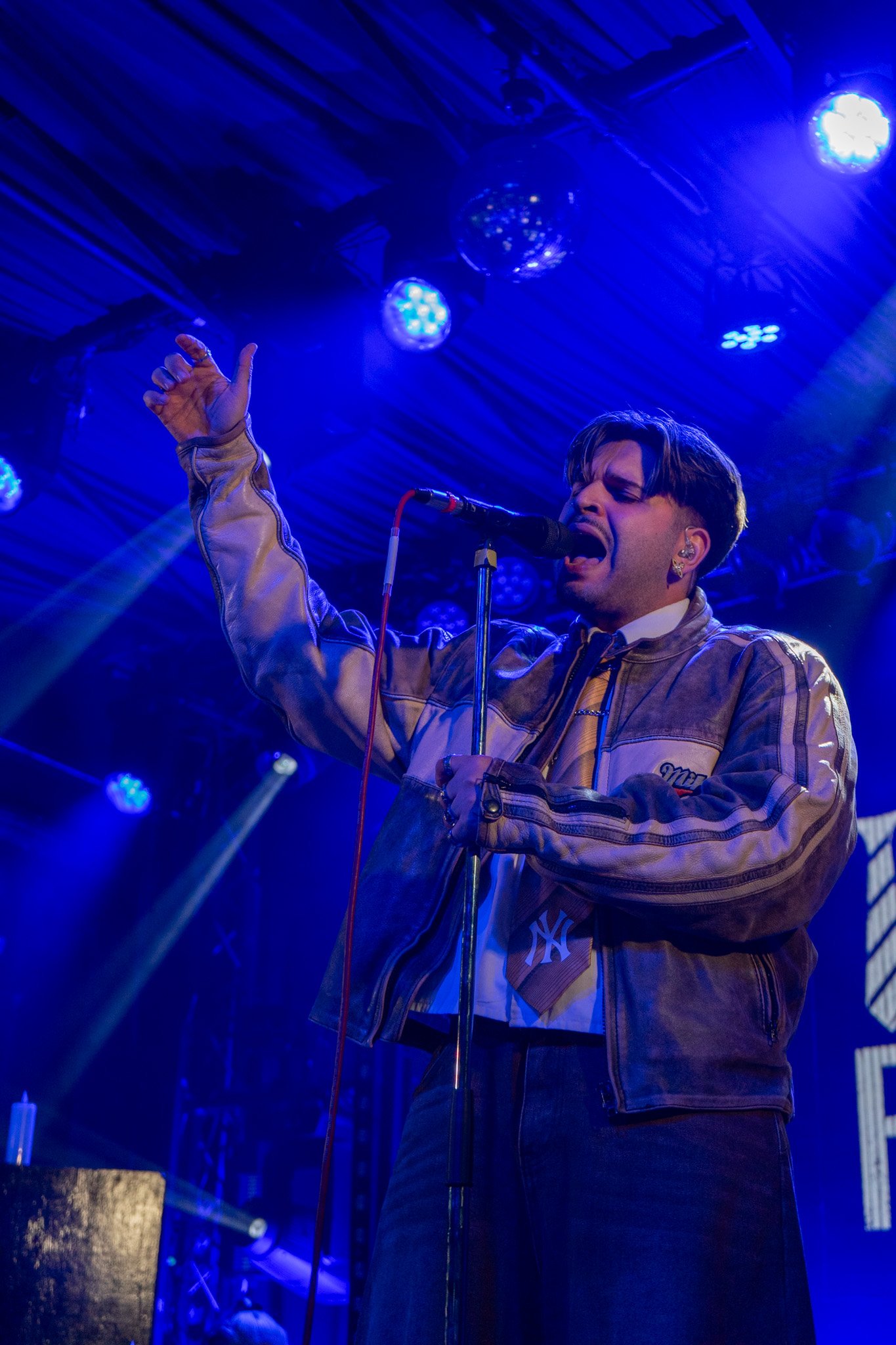 A male singer performs on a stage illuminated with blue stage lights, wearing a beige baseball jersey and passionately singing into a microphone.