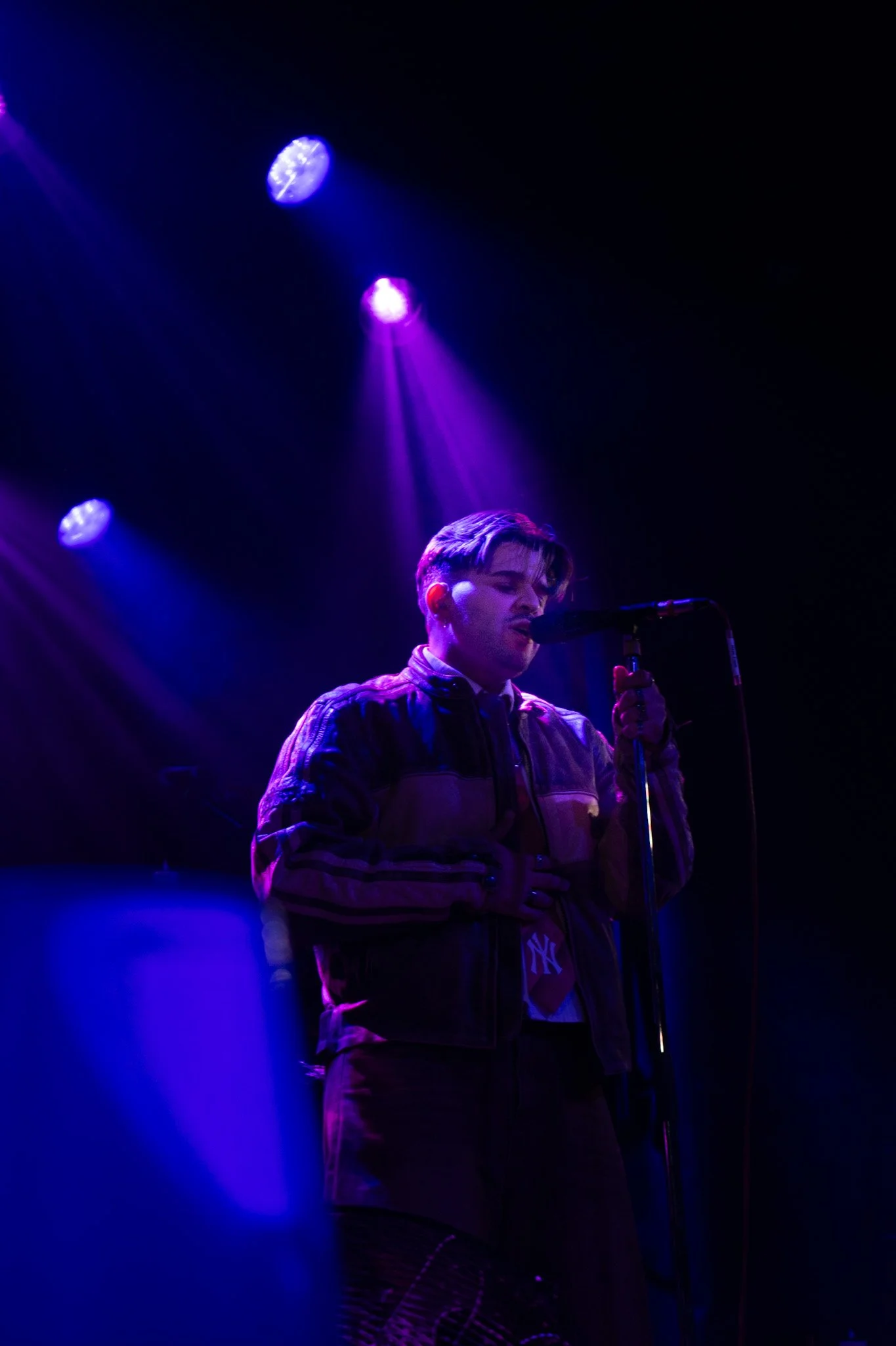 A young man with styled hair singing into a microphone on stage, illuminated by purple and blue stage lights in a dark surroundings.