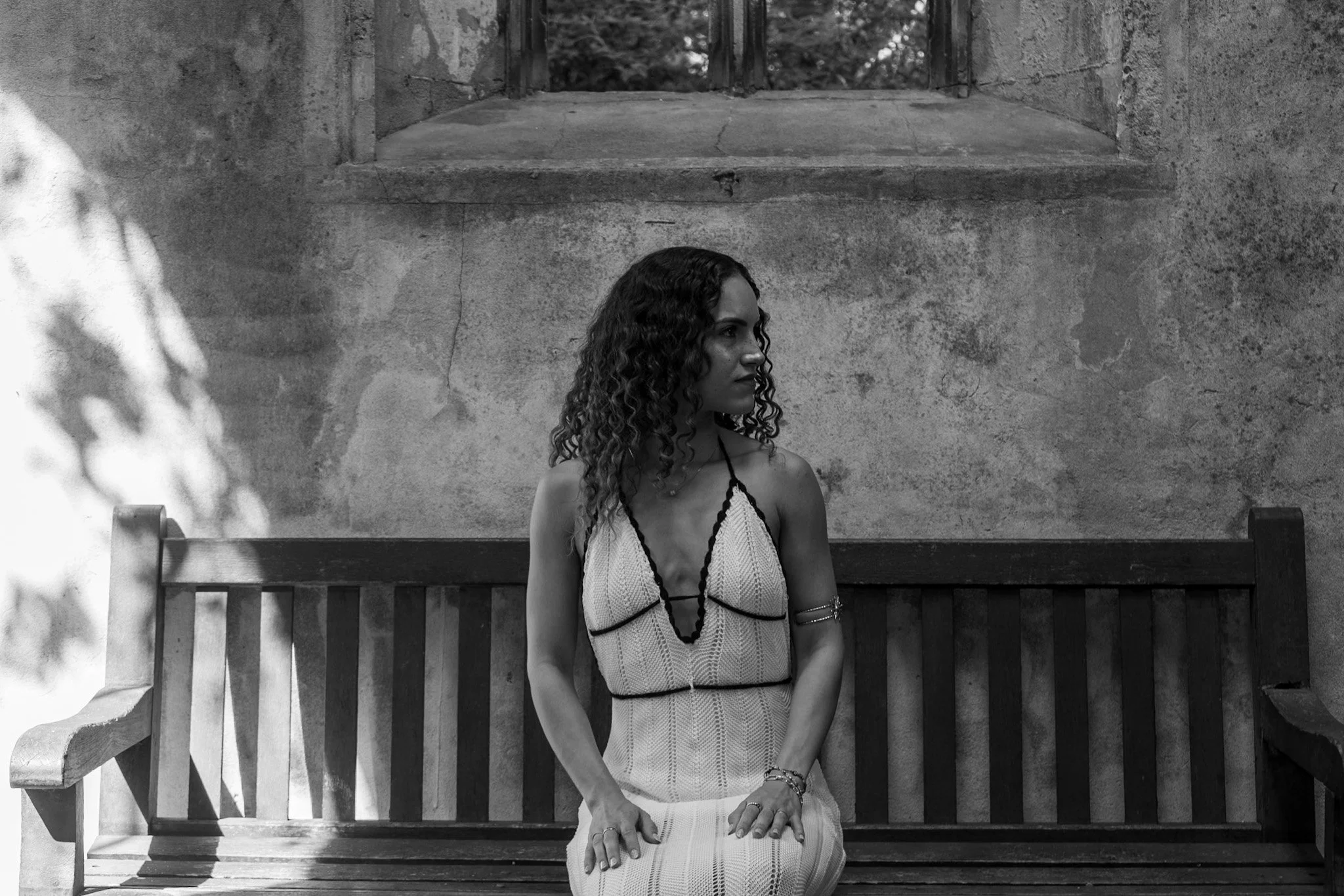 A woman with curly hair sitting on a wooden park bench, wearing a sleeveless striped dress, with a concrete wall behind her and a window above.