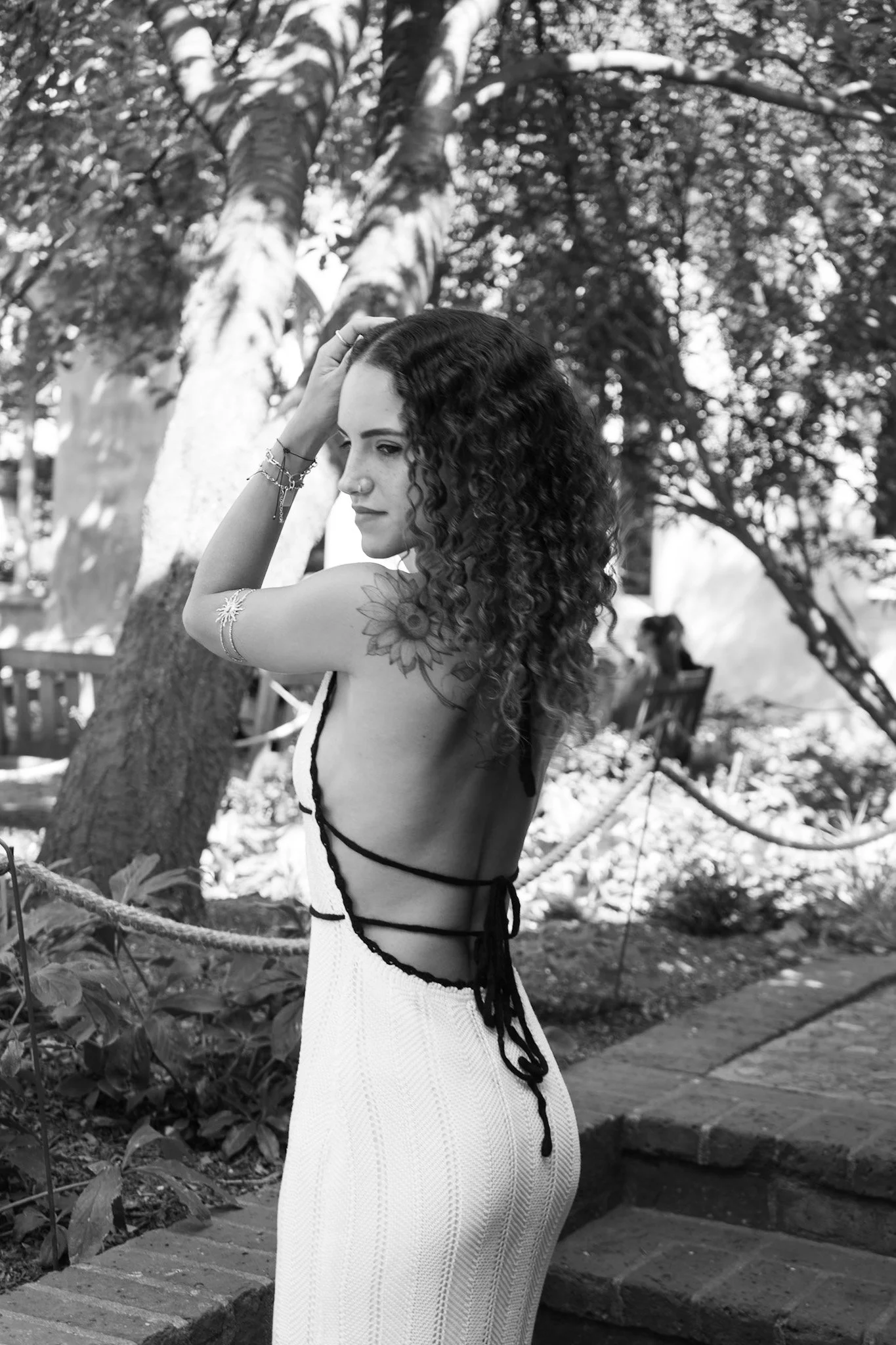 A woman with curly hair and floral tattoo on shoulder, wearing a sleeveless dress with side strings, standing outdoors near a tree and a bench.
