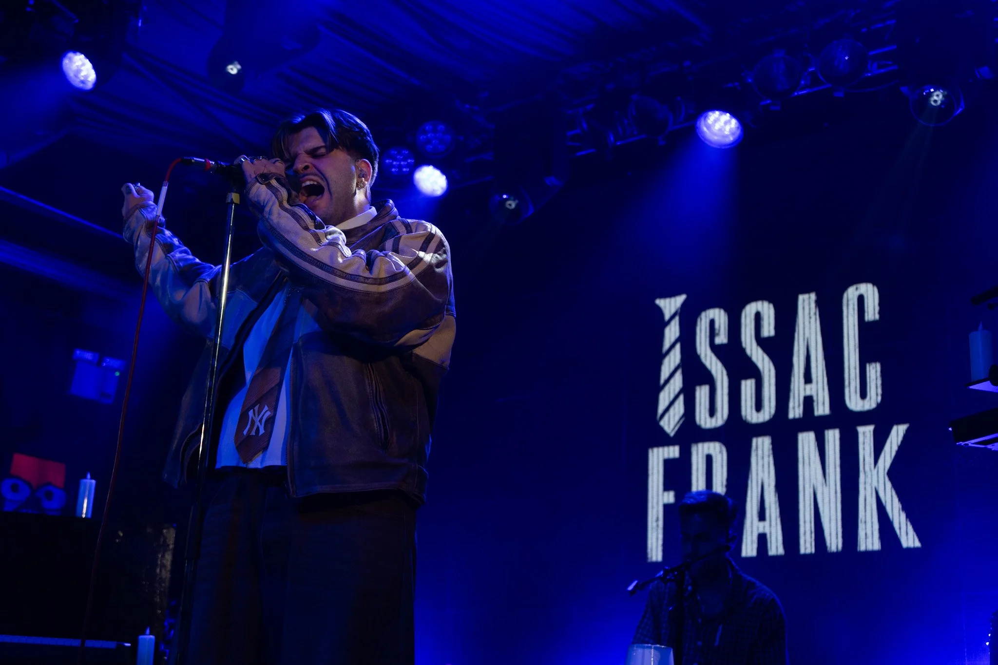 A male singer passionately performs on stage, holding a microphone with both hands, wearing a jacket with the New York Yankees logo, and singing with his eyes closed. The stage is illuminated with blue lights, and a large screen behind him displays p