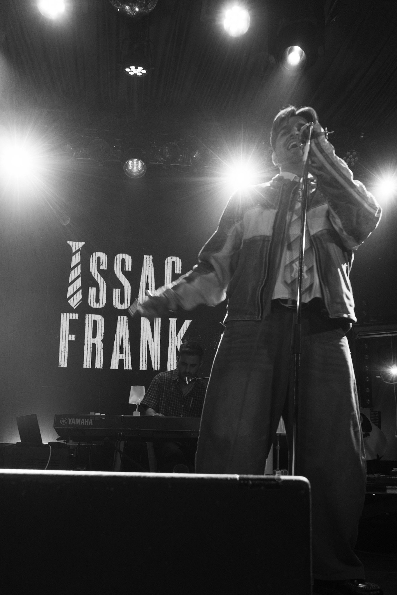 A black and white photo of a man singing into a microphone on stage with bright lights behind him. A keyboardist is visible in the background, playing a Yamaha keyboard. Part of a sign with the text 'ISS' and 'FRANK' is visible behind the performers.