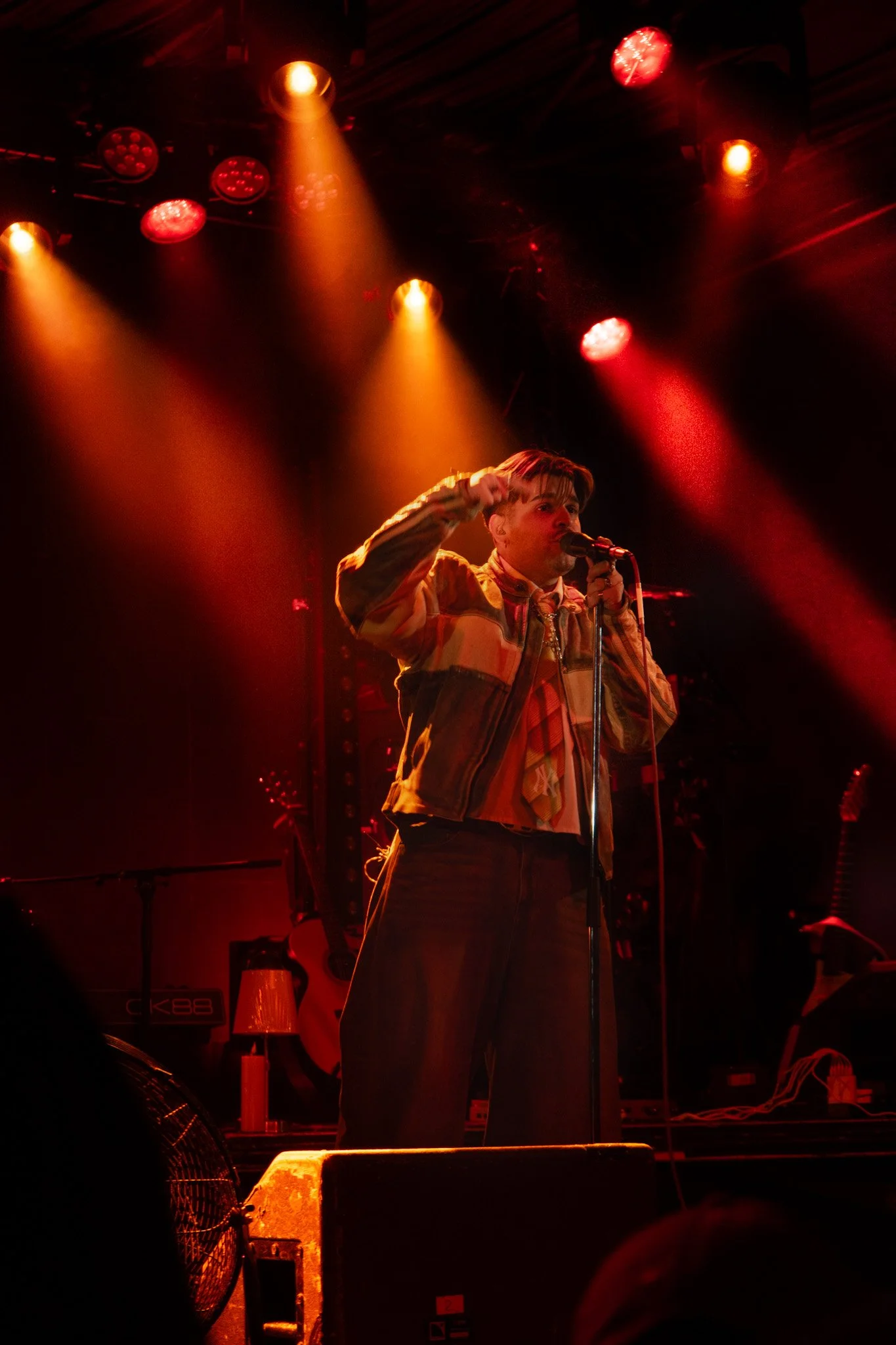 Musician on stage performing with a microphone, under red and yellow stage lights, wearing a jacket and plaid shirt.