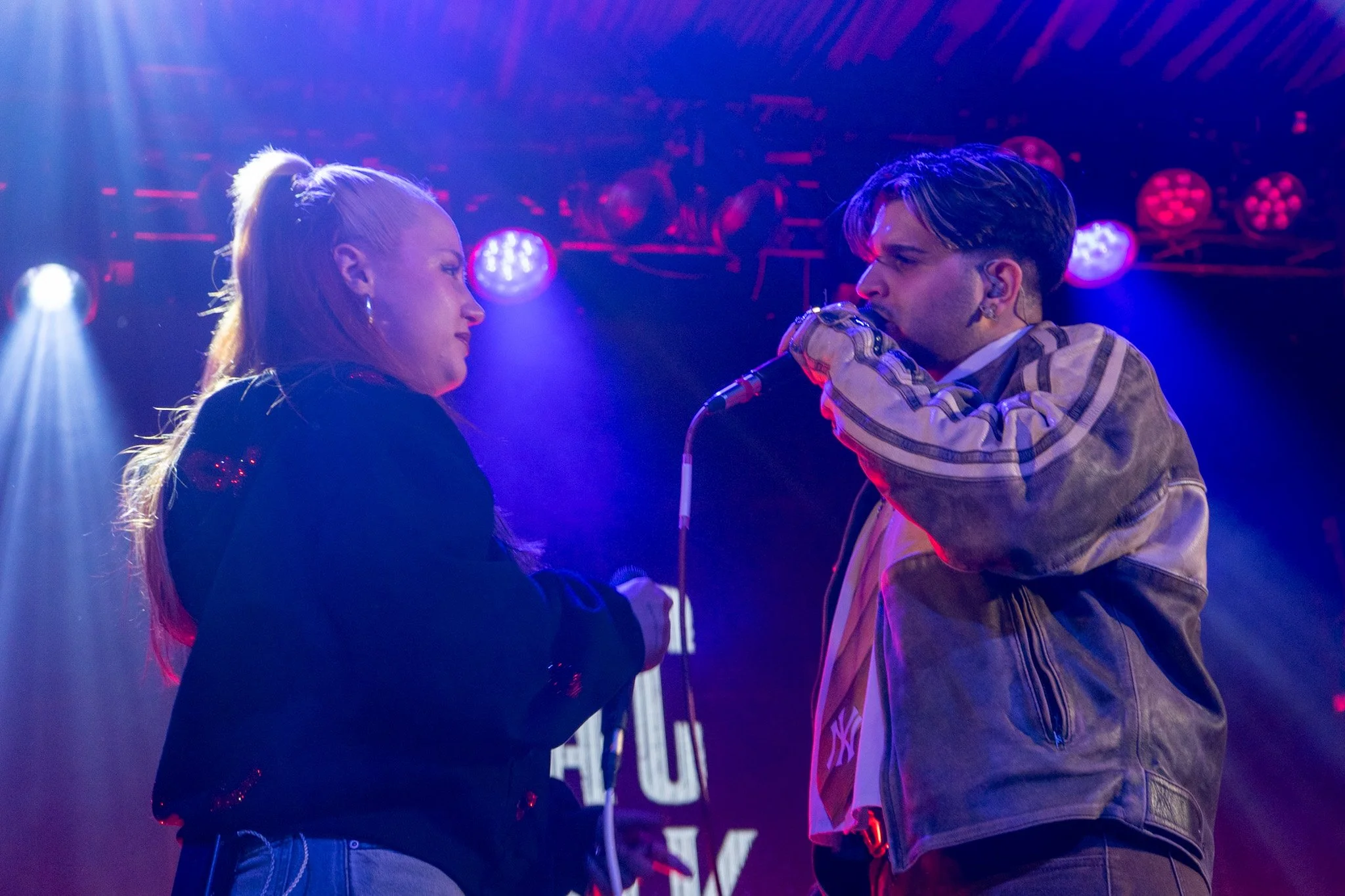 A woman and a man face each other closely on a stage with colorful purple and blue lights, suggesting a performance or intimate moment.