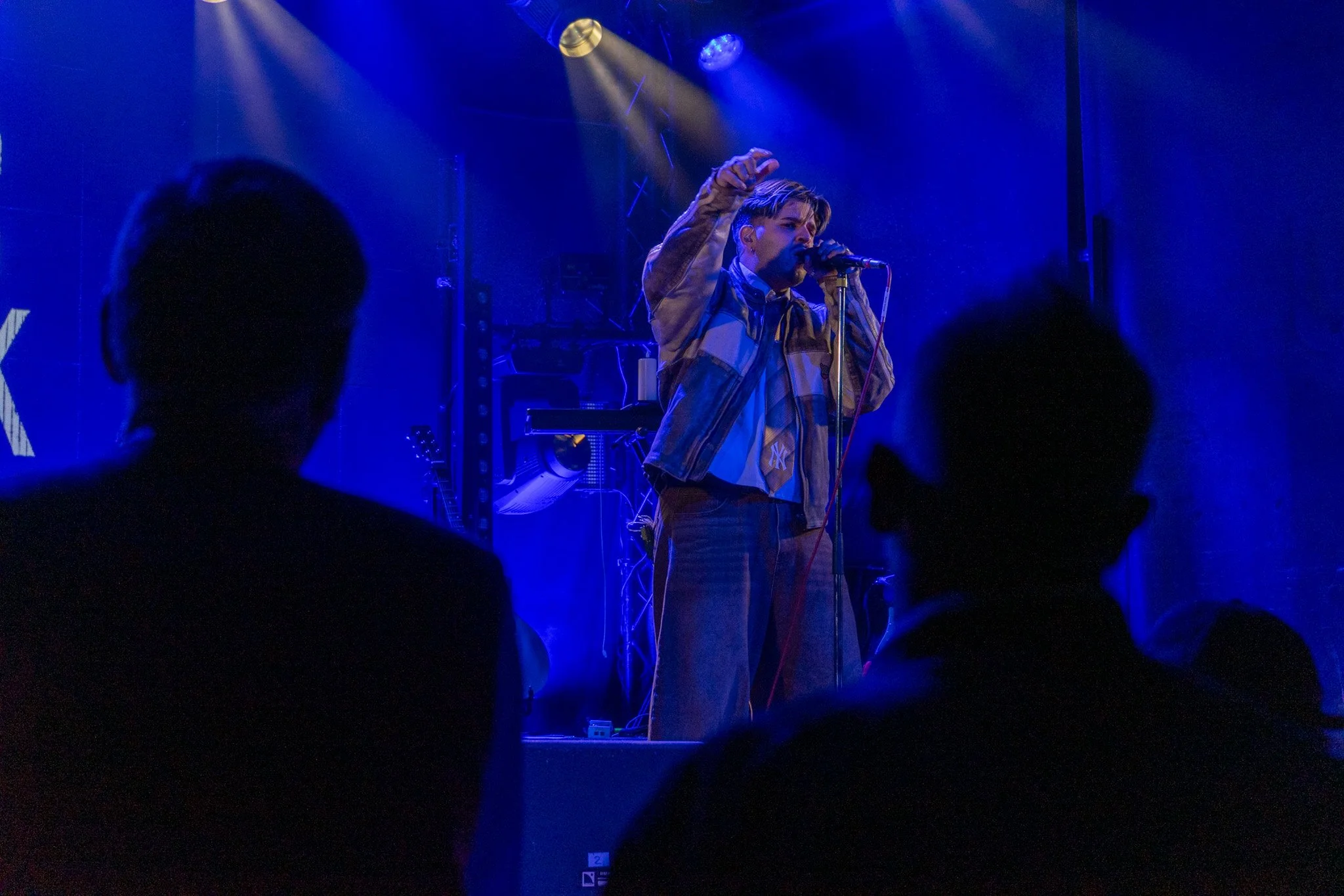 A person performing on stage, singing into a microphone, illuminated by blue and yellow stage lights, with an audience in front.