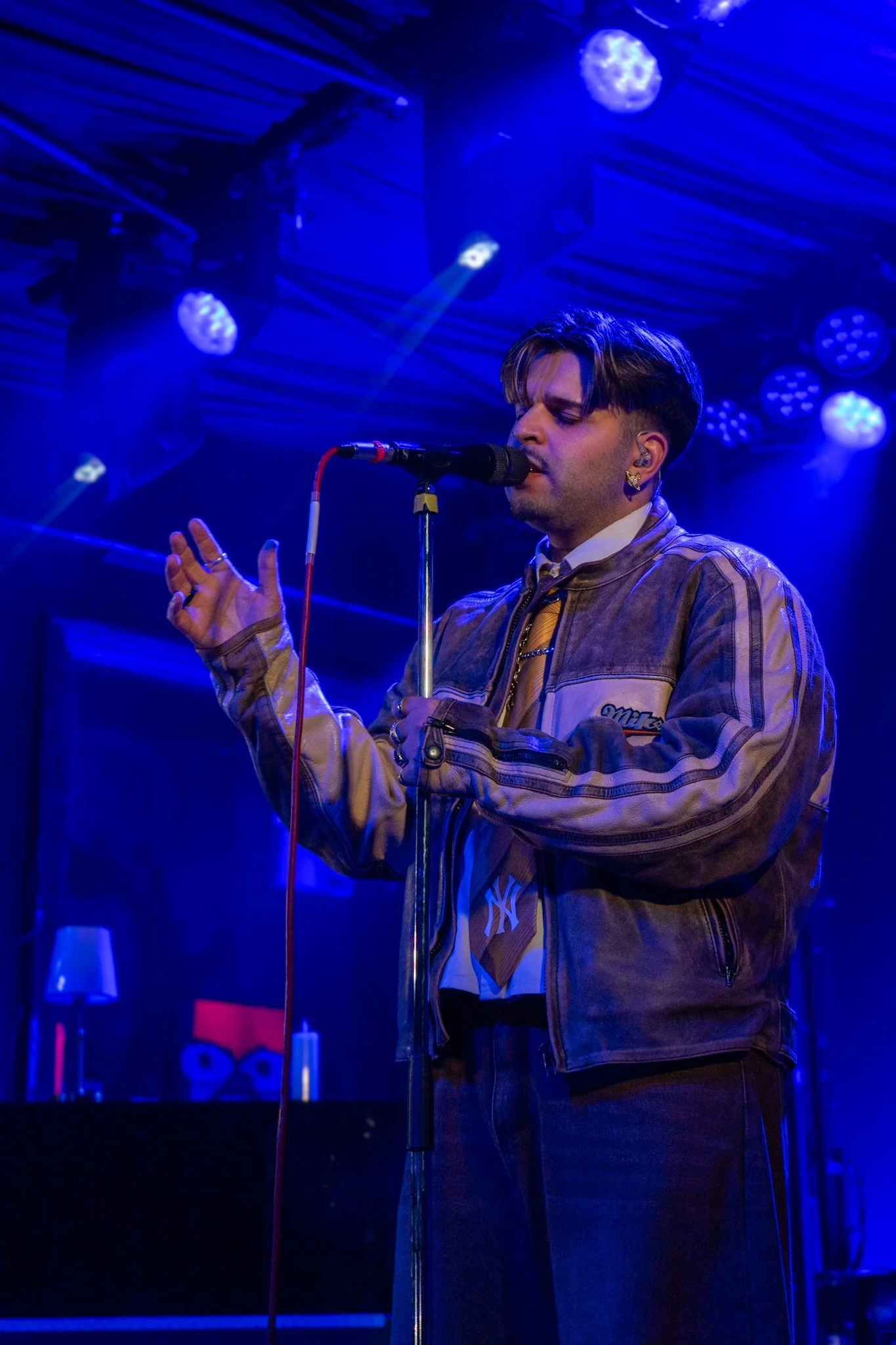 Young man with dark hair singing into a microphone on a stage illuminated by blue lighting, wearing a beige jacket with patches and a yellow tie.