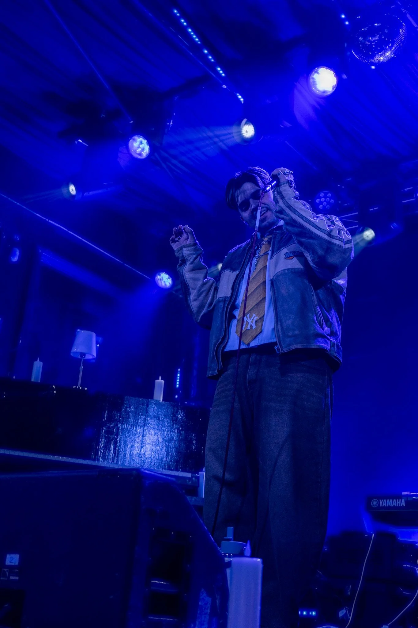 A person wearing a yellow shirt and a beige jacket with a New York Yankees logo, standing on a stage under blue and purple lighting. The person is holding a microphone close to their face.