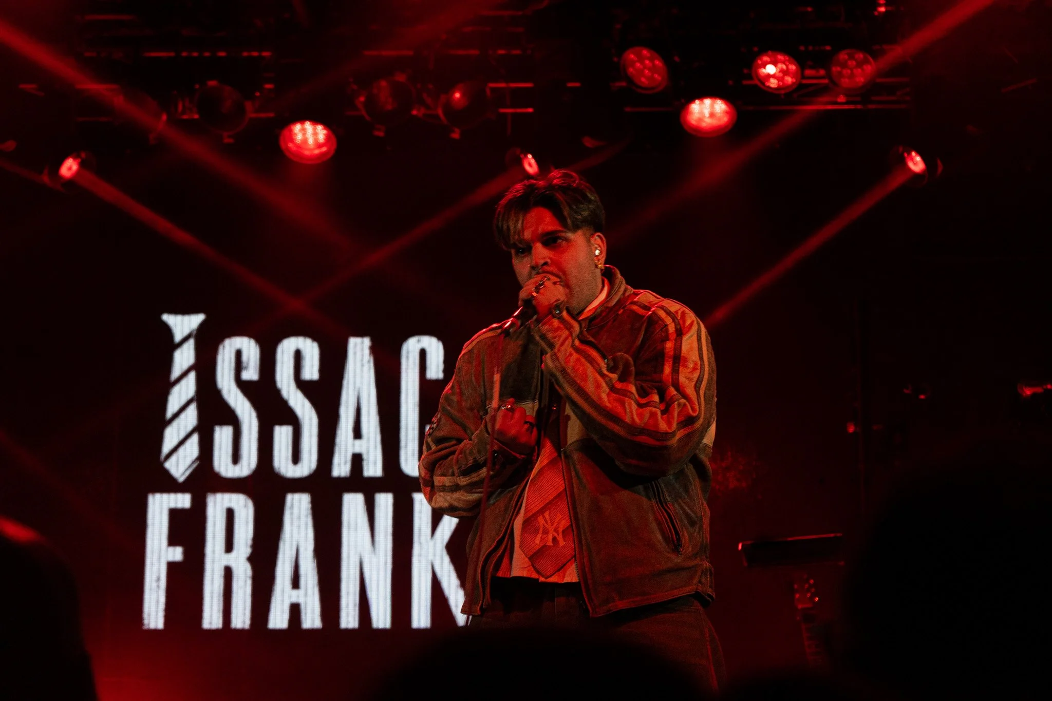 A male singer performing on stage with red lighting and a large screen in the background displaying text.