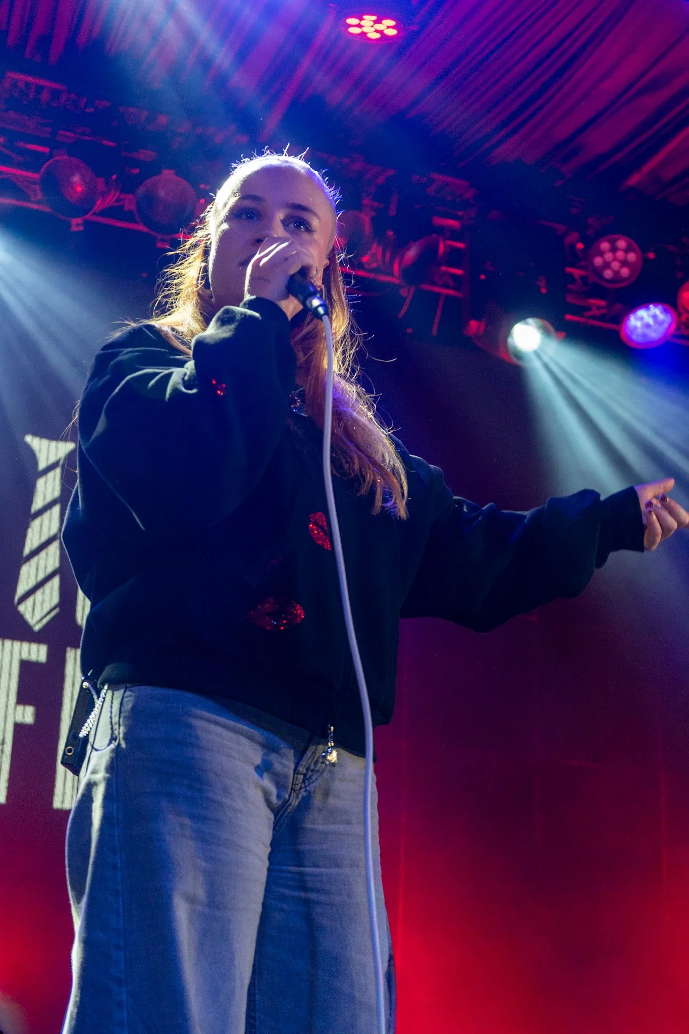 A young woman with long hair holding a microphone on stage with colorful stage lights shining down. She is wearing a black jacket and gray pants.