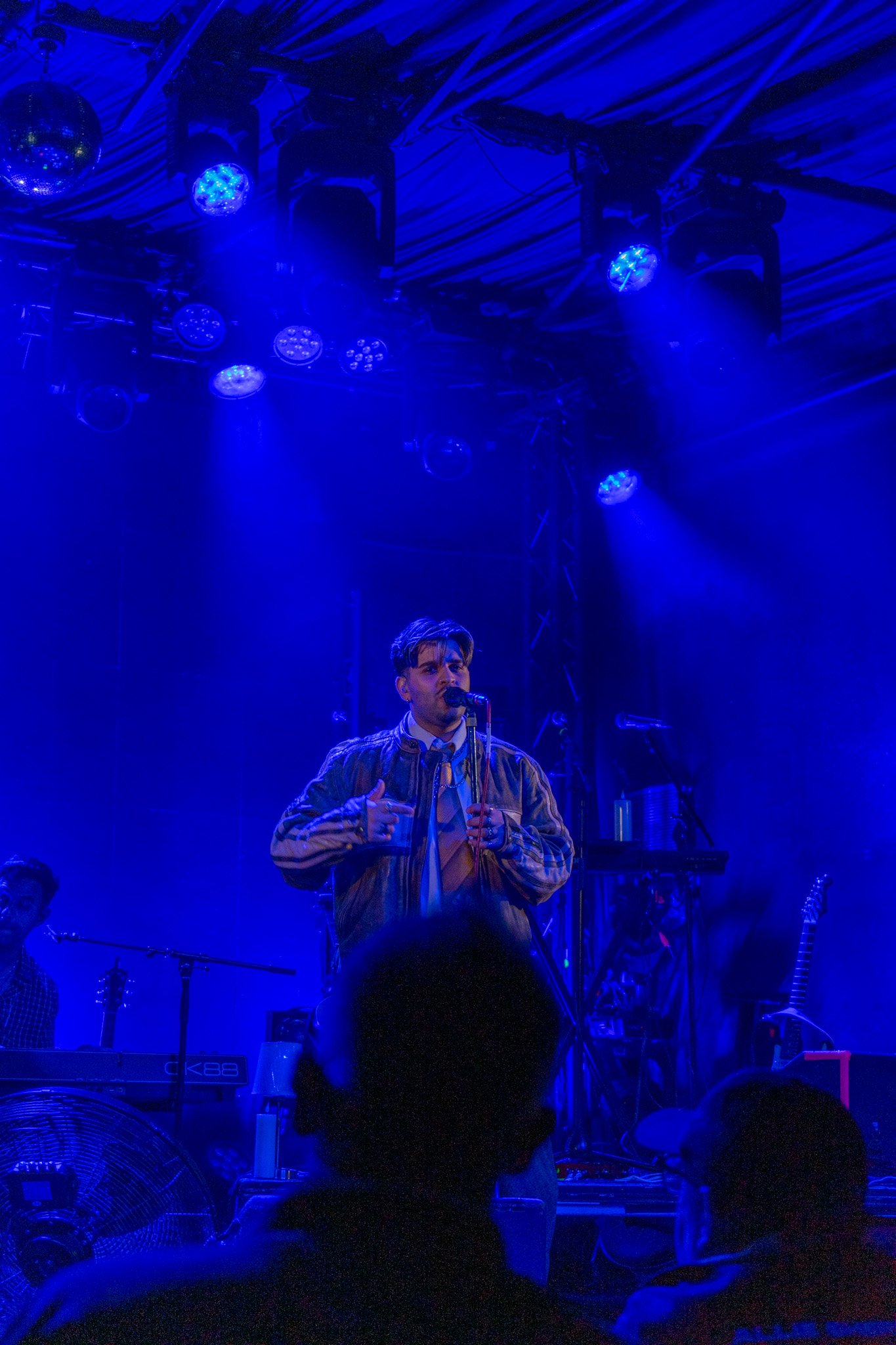 A man singing into a microphone on stage under blue lighting, with musical instruments and equipment visible in the background.