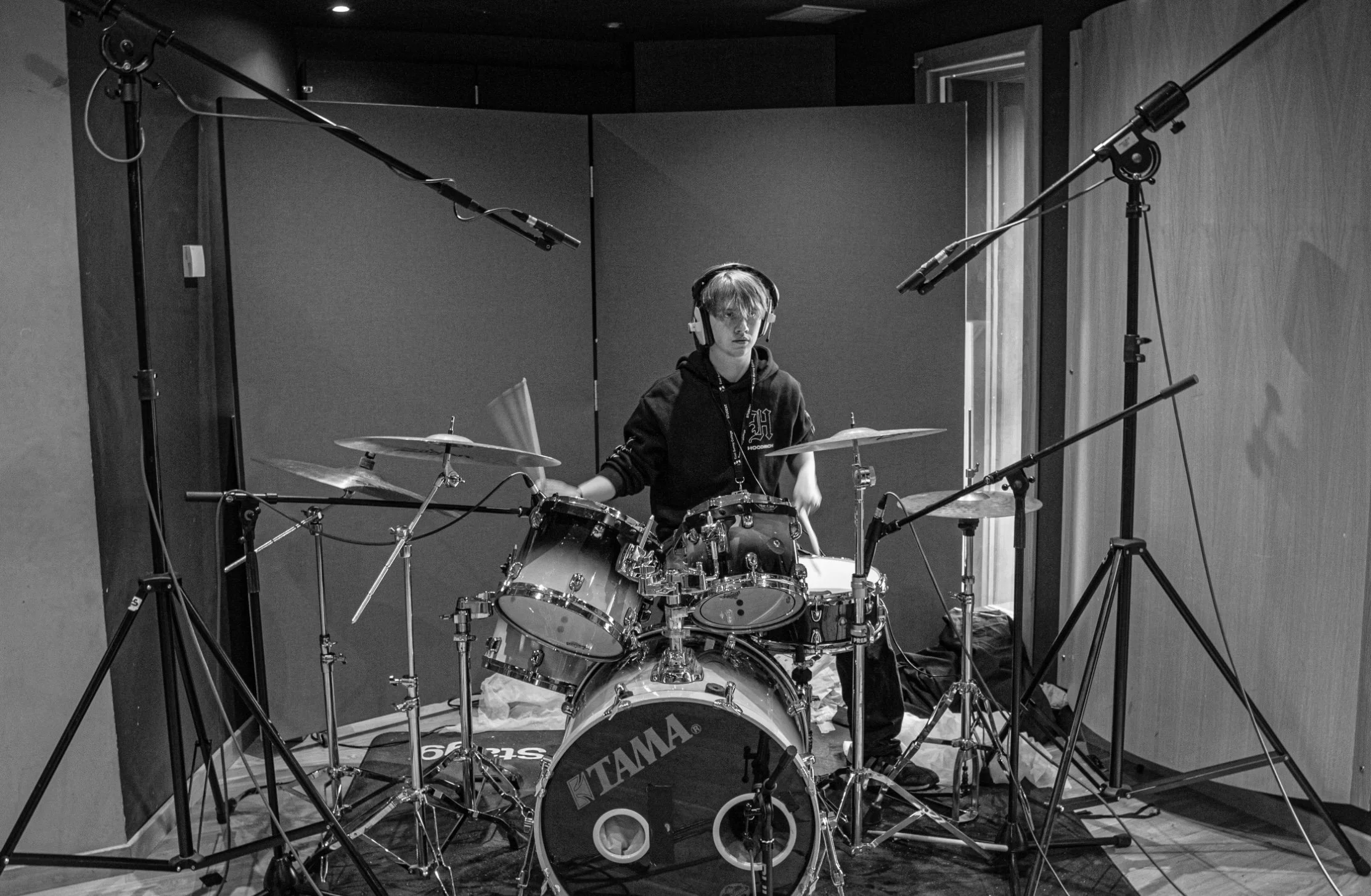 A young person wearing headphones is sitting behind a drum set in a recording studio, surrounded by multiple microphones and soundproofing panels.