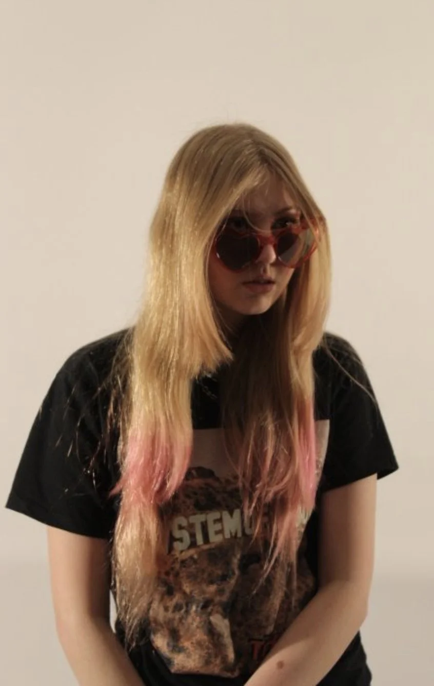 A young woman with long, pink-tinted blonde hair wearing red sunglasses and a black T-shirt with a graphic design.
