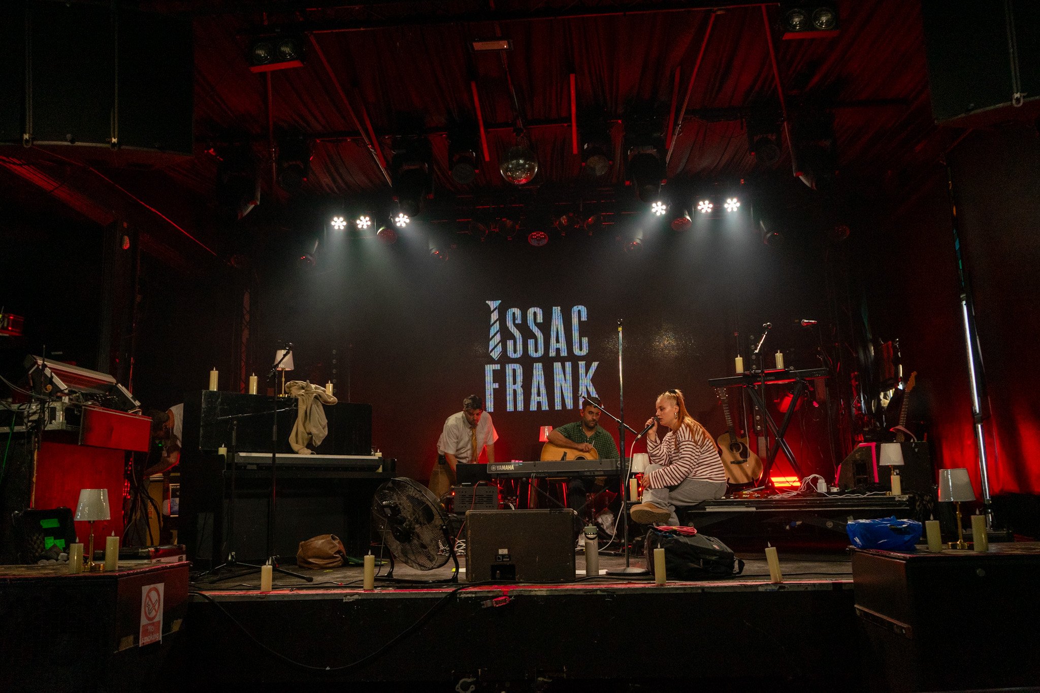 A stage with musicians and equipment, featuring a large screen with the text 'ISSAC FRANK', hanging lights, and various musical instruments, candles, and people preparing for a performance.